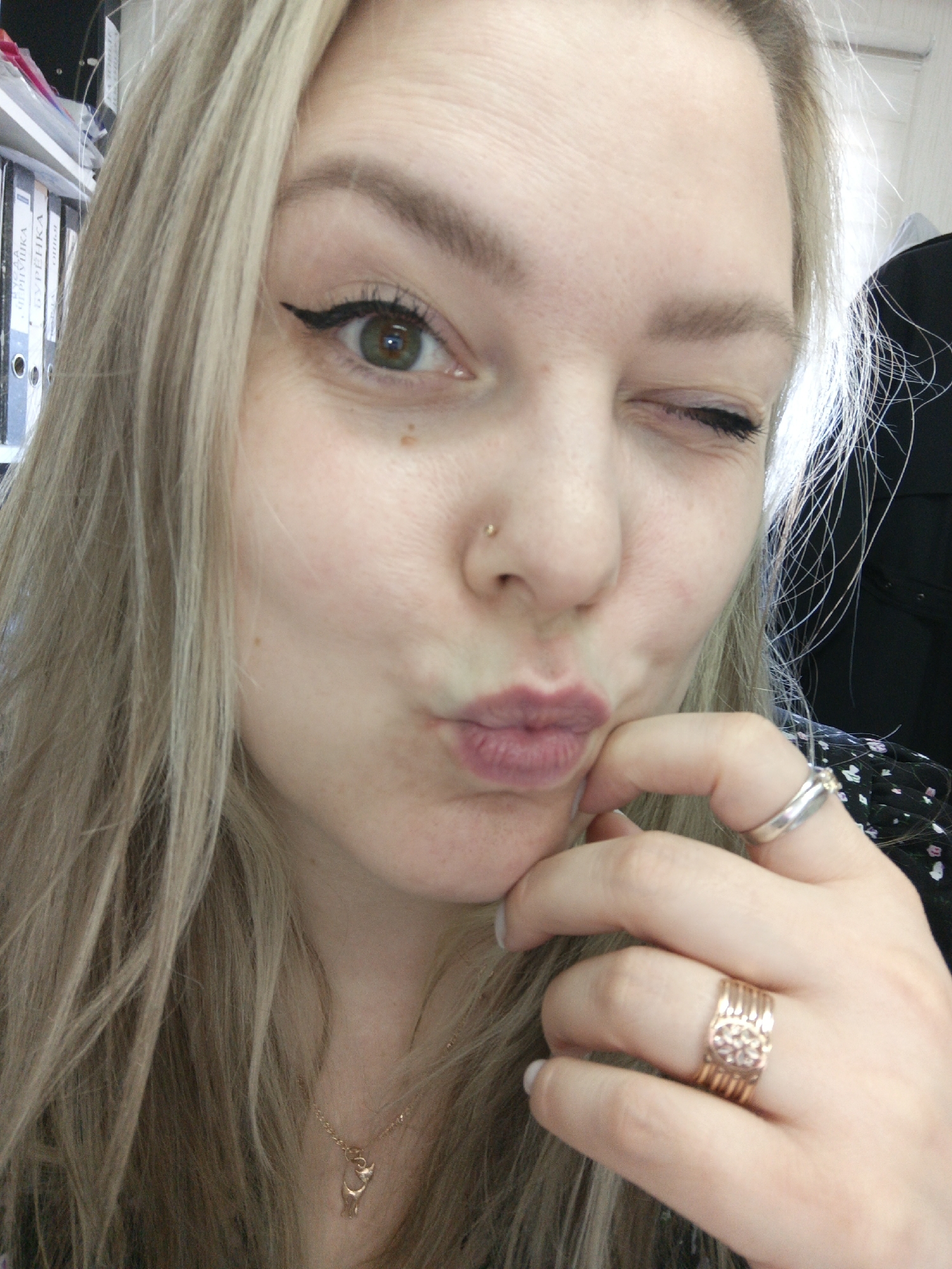 A woman with light blonde hair and fair skin winks at the camera. She has green eyes and is wearing eyeliner. She has a nose piercing and a small gold necklace. She has two rings on her right hand. Her lips are pursed in a kiss. The background appears to be an office setting, with files visible behind her. It is not possible to confirm the location as Noviy, Russia from this image.