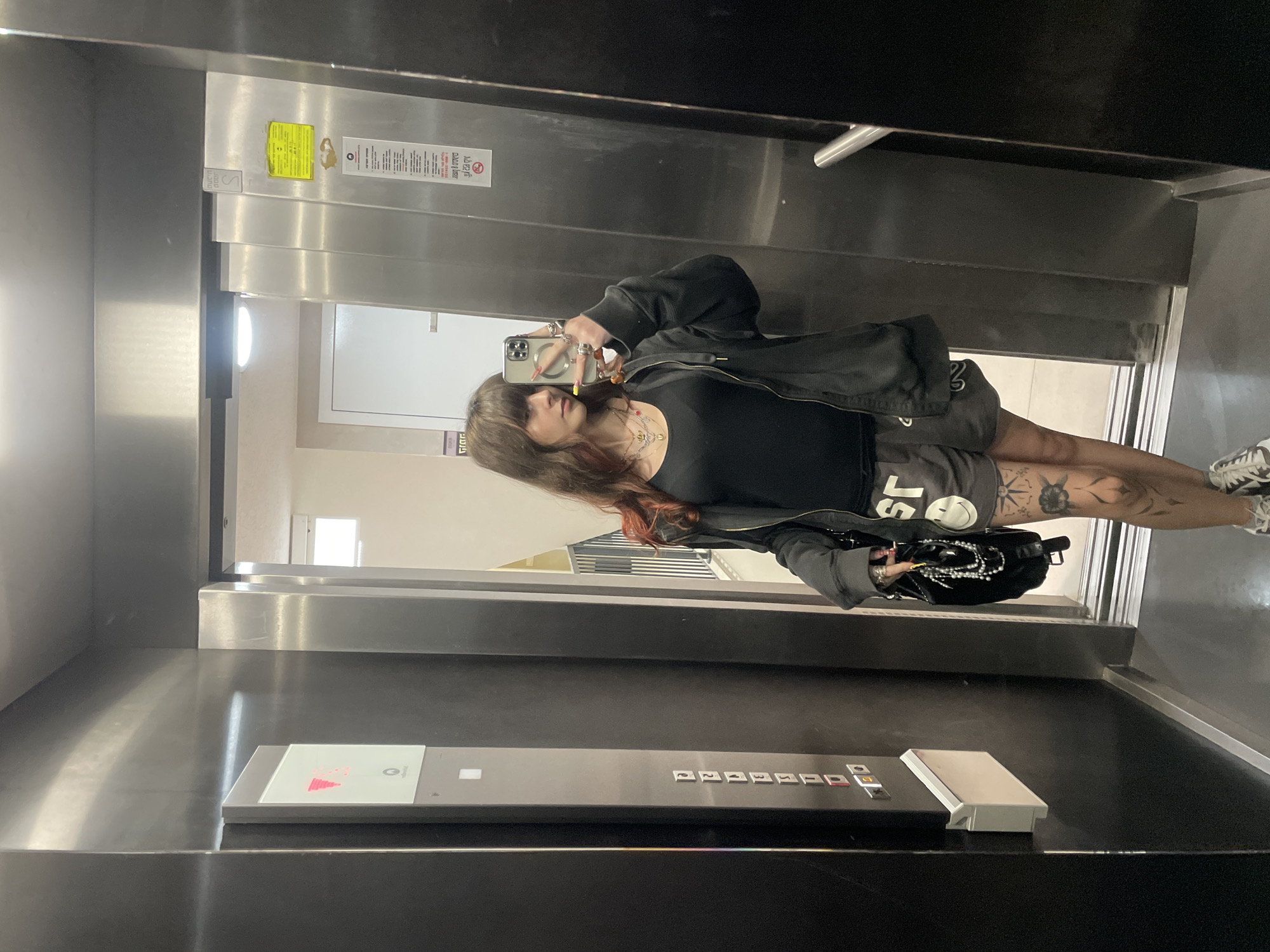 An individual is taking a selfie in an elevator. The elevator has a metallic interior, with a control panel visible featuring floor buttons. The person is wearing a dark top, shorts, and a jacket. Tattoos are visible on their leg. The setting is indoors, presumably within a building in Raanana, Israel, though there is no other information in the image that indicates that.