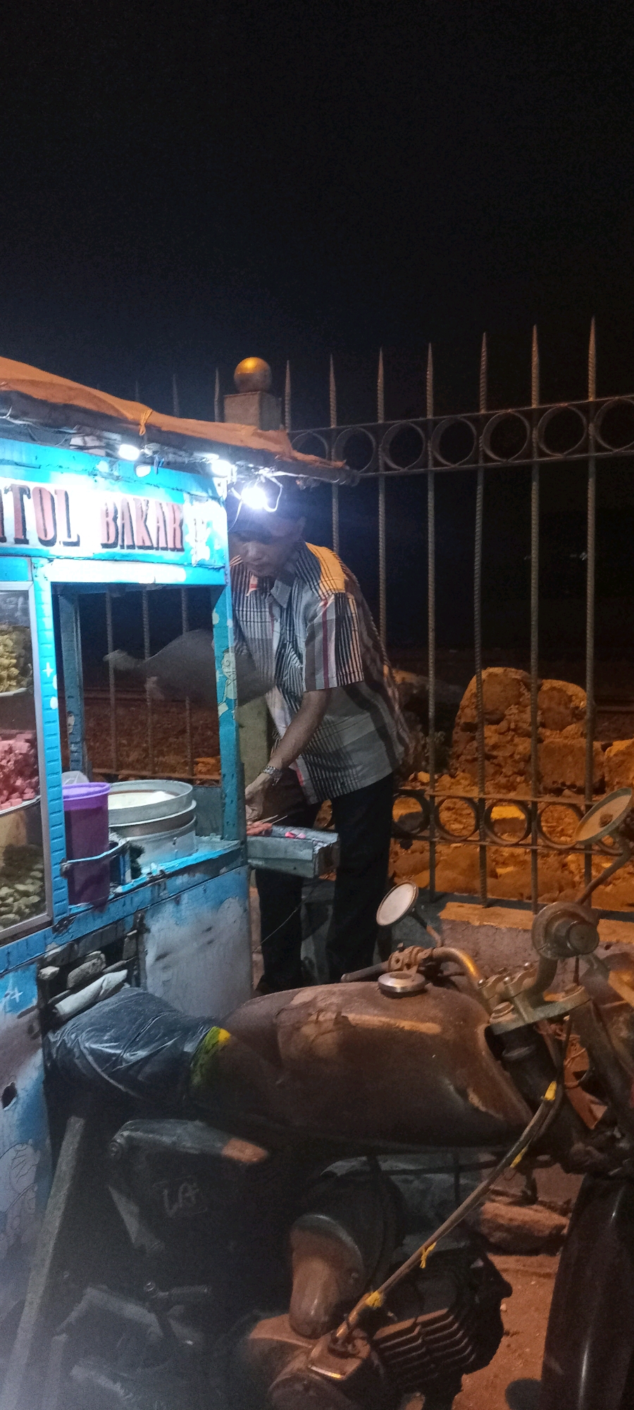The image shows a street food vendor at night in Singogalih, Indonesia, preparing food. A man, possibly the vendor, is wearing a striped shirt and a headlamp, and is standing next to a food cart. The cart has blue trim and a sign that reads "TOL BAKAR". On the cart, there are containers of food and what appears to be a grill with skewers of food. In the foreground, part of a motorcycle is visible. The background shows a metal fence and some stone wall. The lighting is dim, suggesting it is nighttime.