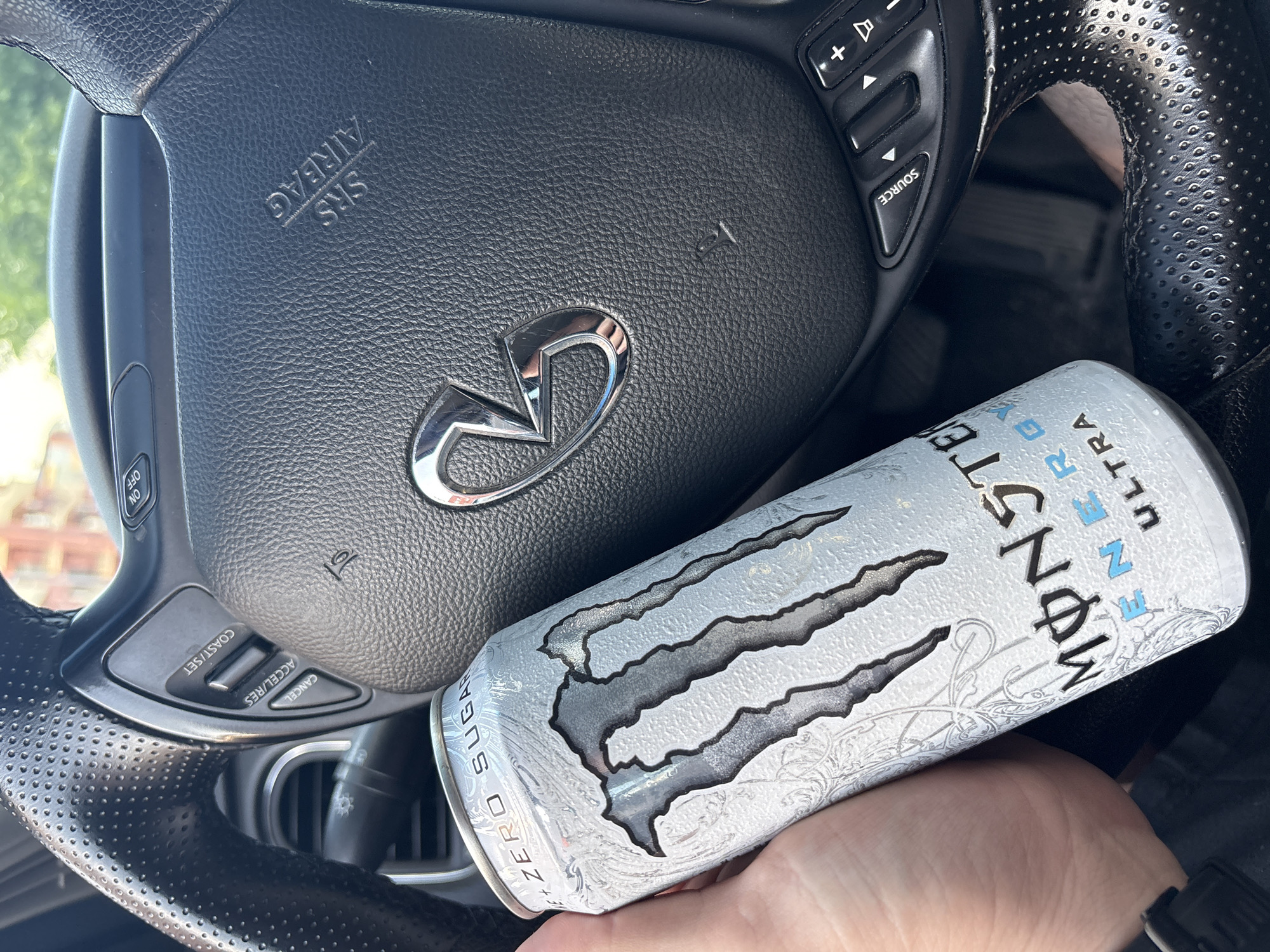 An eye-level shot inside a car, focusing on a hand holding a Monster Ultra energy drink can, with a steering wheel dominating the background. The scene appears to be set in Plovdiv, Bulgaria, under bright daylight conditions.
The main subjects are a person's hand holding a white and silver Monster Ultra energy drink can, and a black leather steering wheel. The steering wheel, likely belonging to an Infiniti vehicle as suggested by the prominent chrome logo in the center, features various control buttons. These include audio controls with "+" and "-" for volume and a "SOURCE" button, as well as cruise control settings labeled "COAST/SET", "ACCEL/RES", and "CANCEL". Additional buttons for "ON" and "OFF" are visible, possibly related to cruise control or other functions. The text "SRS AIRBAG" is embossed on the steering wheel's central pad, indicating the presence of an airbag. Parts of the steering wheel have a perforated leather texture.
The Monster Energy can is clearly visible, showcasing its distinctive claw-mark logo in silver against a white background with subtle patterns. Text on the can reads "MONSTER ENERGY ULTRA" and "ZERO SUGAR". The activity depicted is simply holding the energy drink within the car environment, possibly while driving or parked. No other people are visible, and emotions cannot be discerned.