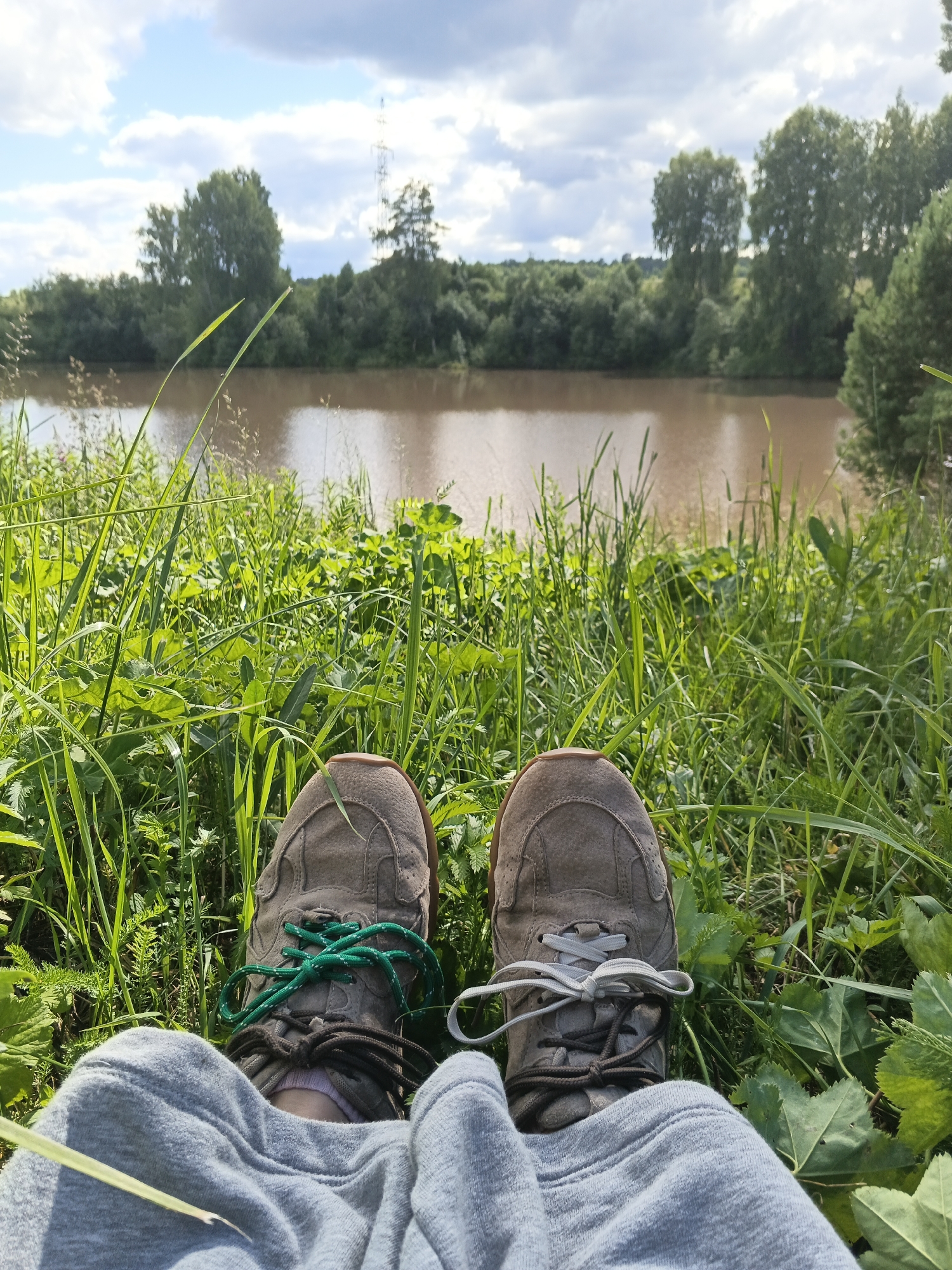 The image depicts a first-person perspective of a person sitting in lush green grass by a muddy river. The subjects are the person's feet, clad in gray sneakers with contrasting green and gray shoelaces, and their gray sweatpants. The scene is a natural landscape with a wide river in the middle ground, its surface reflecting the sky and surrounding trees. The background is a dense line of green trees, and further in the distance, a tall electrical tower is visible against a partly cloudy sky. The overall setting suggests a peaceful outdoor environment, likely a rural or semi-rural area, consistent with the Medvedevka, Russia location. The bright sunlight and clear sky, interspersed with clouds, indicate it is daytime, likely late morning or afternoon. The abundance of green grass and foliage suggests a warm season, possibly spring or summer. There are no visible text elements in the image. The composition, with the feet prominent in the foreground and the serene landscape beyond, evokes a sense of relaxation and enjoyment of nature.