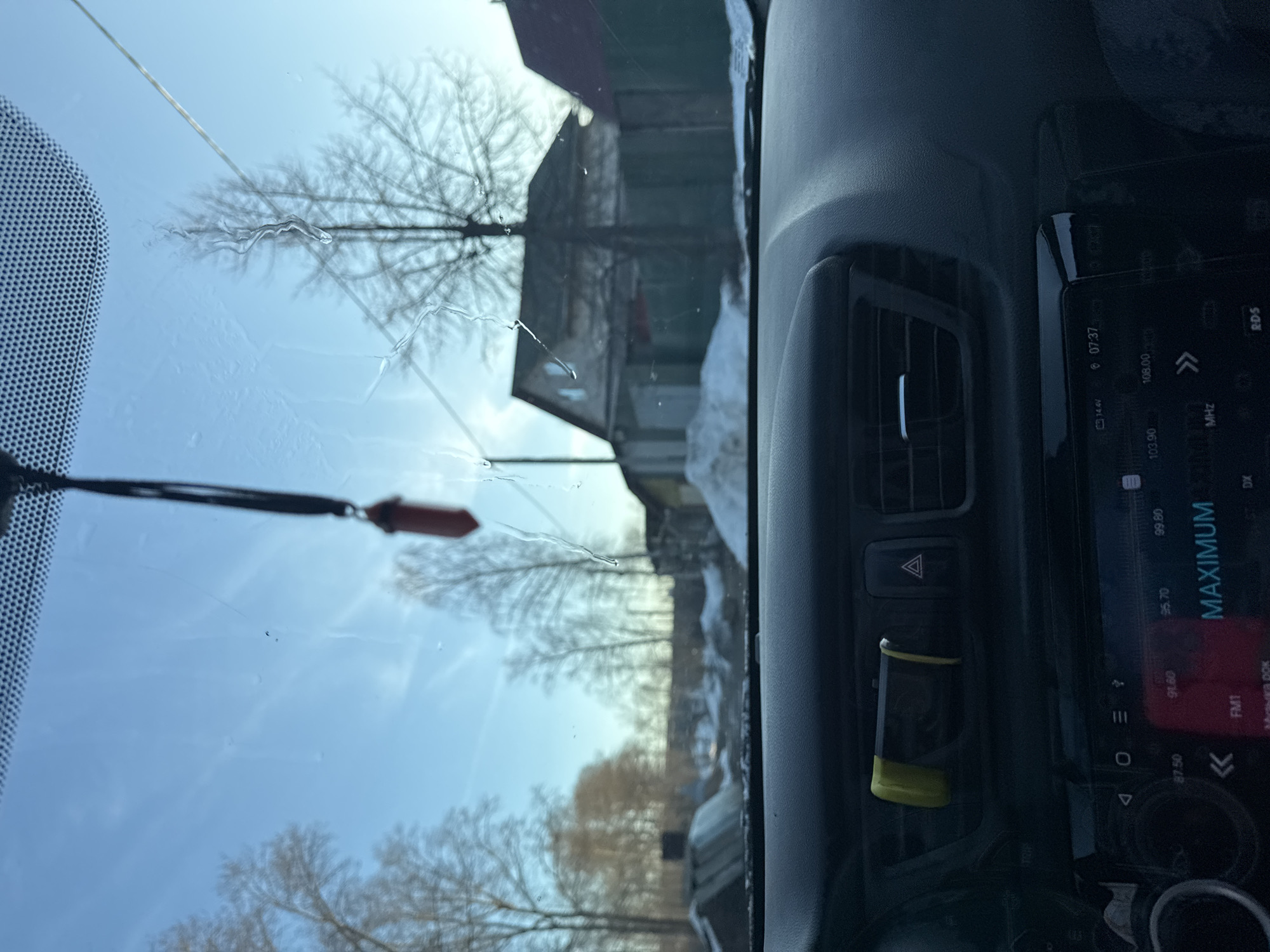 The image appears to be taken from inside a car in Chaikovsky, Russia. The foreground features the car's dashboard, including an air vent, hazard light button, a small accessory (possibly an air freshener or phone holder), and an in-car entertainment system displaying radio frequencies and the word "MAXIMUM". The windshield has a crack and some water spots. The view through the windshield shows a blurry landscape of buildings and bare trees under a bright sky, with utility lines visible. Snow or ice may be present, given the season and location.