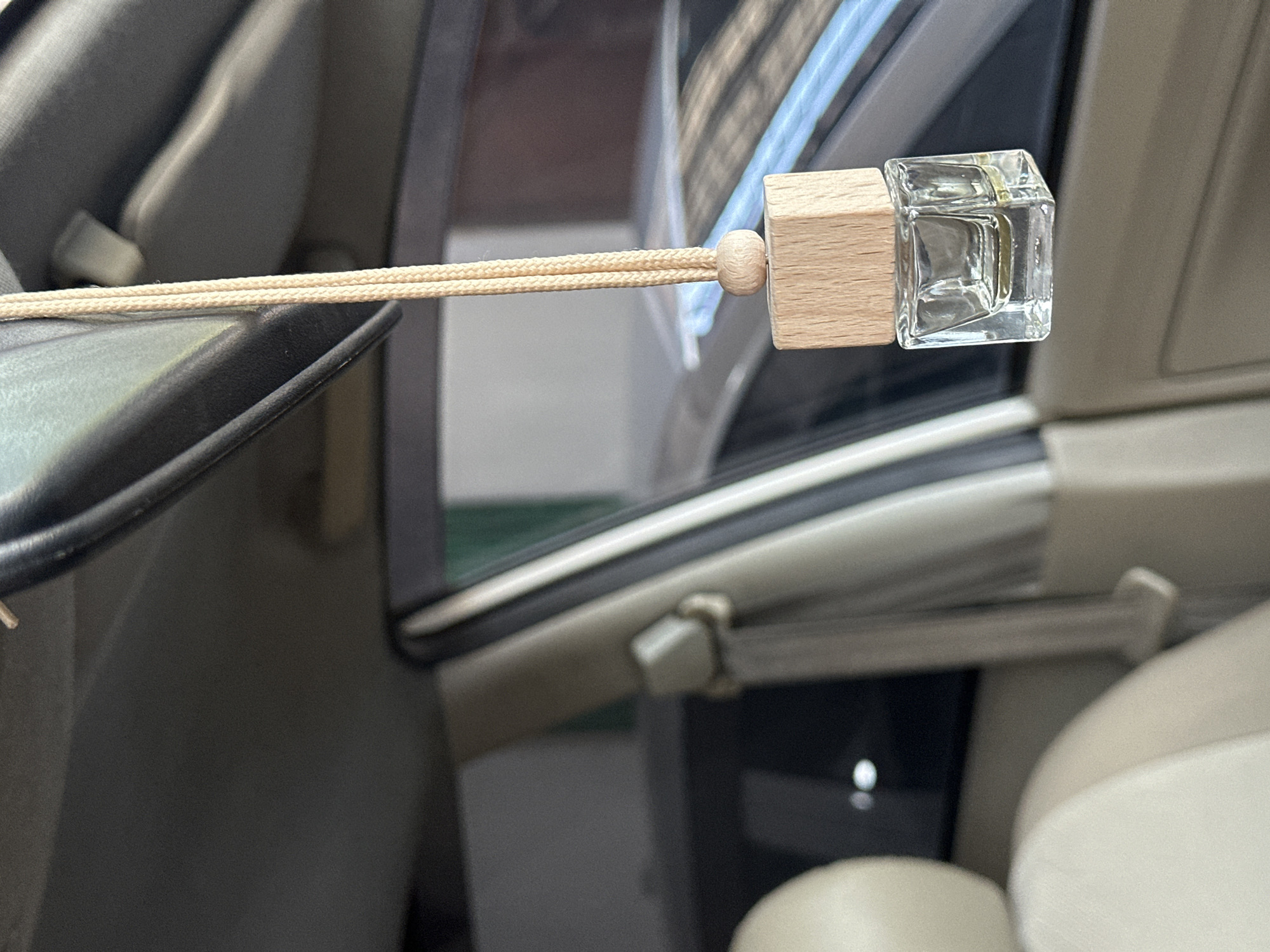 A close-up, eye-level, indoor shot shows a car air freshener hanging from the rearview mirror. The air freshener is a small glass bottle filled with a light-colored liquid, topped with a wooden cap. A thin rope suspends it, adorned with a wooden bead just above the cap. The interior of a car is visible, with a tan seat and seatbelt in the foreground. The background is blurred, showing reflections of buildings and cars, suggesting an urban setting like Bulakrejo, Indonesia. The lighting is natural, coming from outside the car.