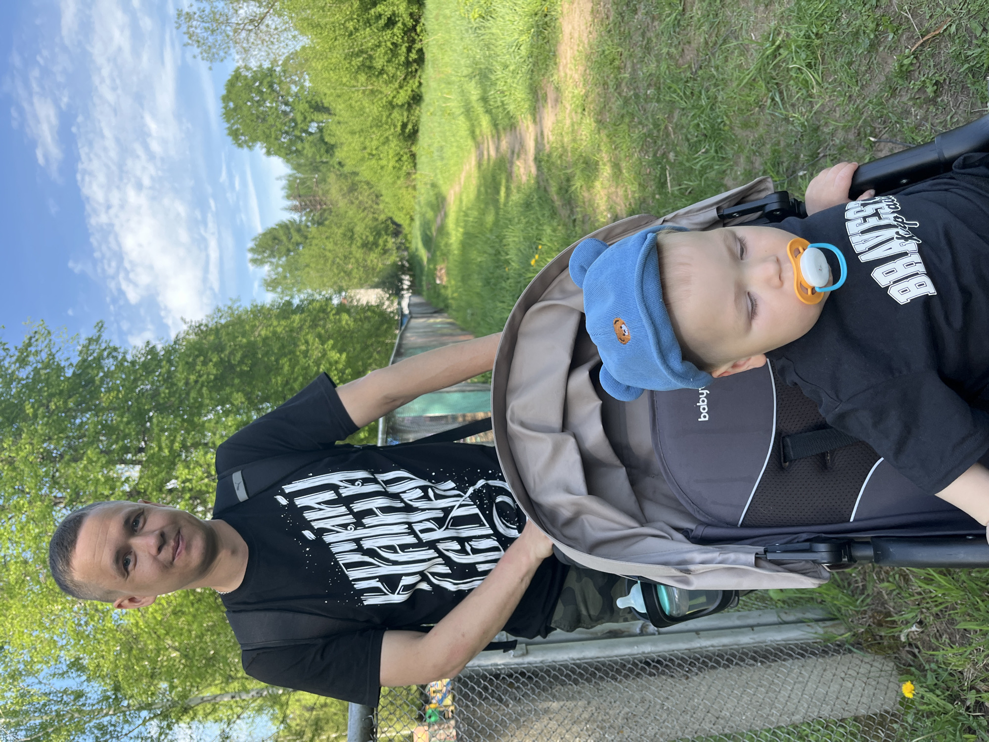 A medium, eye-level, outdoor shot depicts a man pushing a baby stroller on a path near what appears to be a park in Chaikovsky, Russia. The man has a short haircut and is wearing a black T-shirt with a white graphic design. He is looking toward the camera with a faint smile. He has a dark backpack slung over his shoulder. The stroller contains a baby who is asleep. The baby has a blue cap with a bear logo on it and is wearing a black T-shirt. The baby has a pacifier in its mouth. The stroller has a neutral tan canopy. The path is green and grassy, flanked by trees. The background has a glimpse of a partly cloudy sky and more foliage. A chain-link fence is visible in the foreground. The image is rotated 90 degrees clockwise.