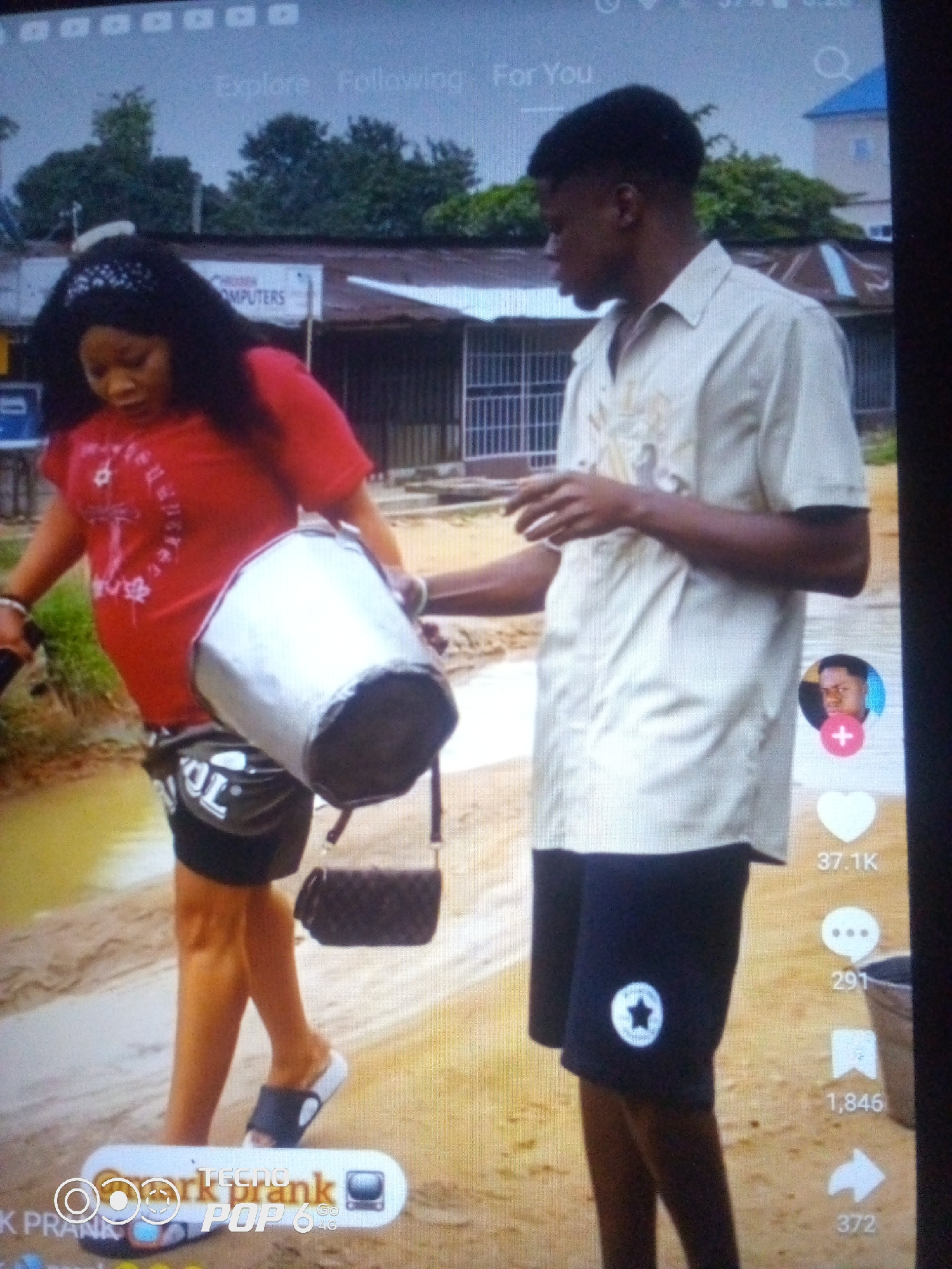 The image depicts a scene on a dirt road, possibly in the city of Howayi, Nigeria, with a few modest buildings in the background. A woman, wearing a red t-shirt and black shorts, is carrying a large, silver cylindrical object that appears to be a container. She has a black headband on and a small, dark purse slung over her shoulder. A young man, dressed in a light-colored collared shirt and dark shorts, stands beside her, holding a phone in his left hand and appears to be interacting with her or the container. The ground is sandy and uneven, with some patchy green vegetation visible on the left side of the frame. There are also icons and text overlaying the image, suggesting it is a screenshot from a social media platform.