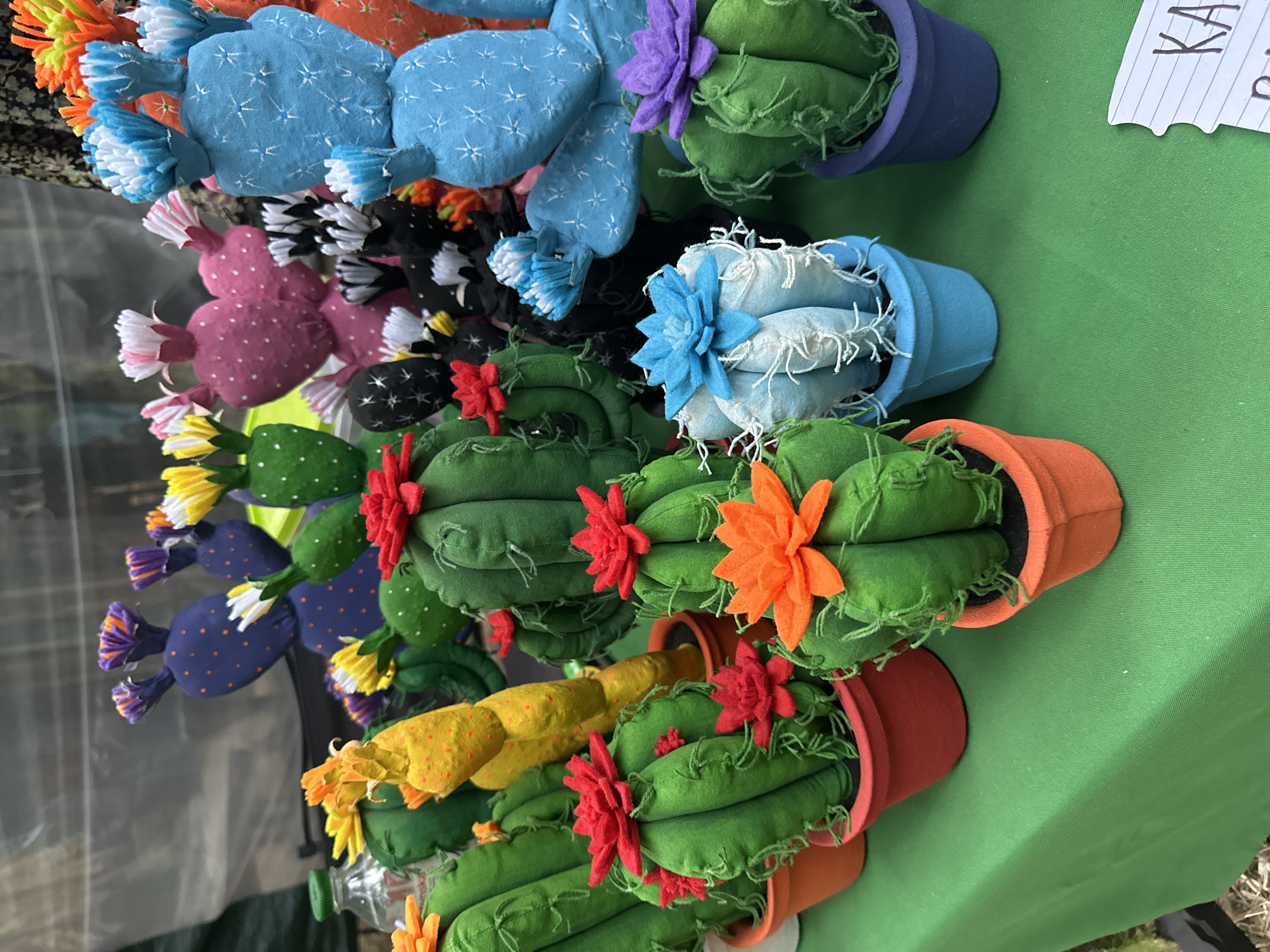 The image displays a collection of handmade fabric cacti arranged on a green tablecloth. The cacti vary in size, shape, and color, with some featuring decorative felt flowers on top. They are potted in colorful fabric pots. In the background, more cacti are visible, along with what appears to be a clear plastic enclosure and a floral patterned fabric. A small white tag with handwritten text is partially visible in the lower right corner. The scene suggests an outdoor market or craft fair, possibly in Kyiv, Ukraine, given the context of a prior request. There are no people clearly visible in this particular frame.