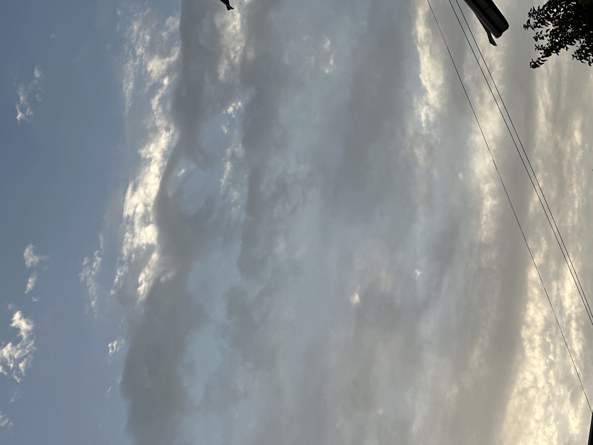 The image depicts a sky filled with clouds, with a portion of a roofline and tree branches visible in the lower right corner. There are also power lines crossing the lower part of the frame. The sky is a mix of blue and gray clouds, with some areas illuminated by sunlight. No people or specific actions are observable. The image does not contain enough visual information to determine its location within the city of Sasbaga, Uzbekistan.