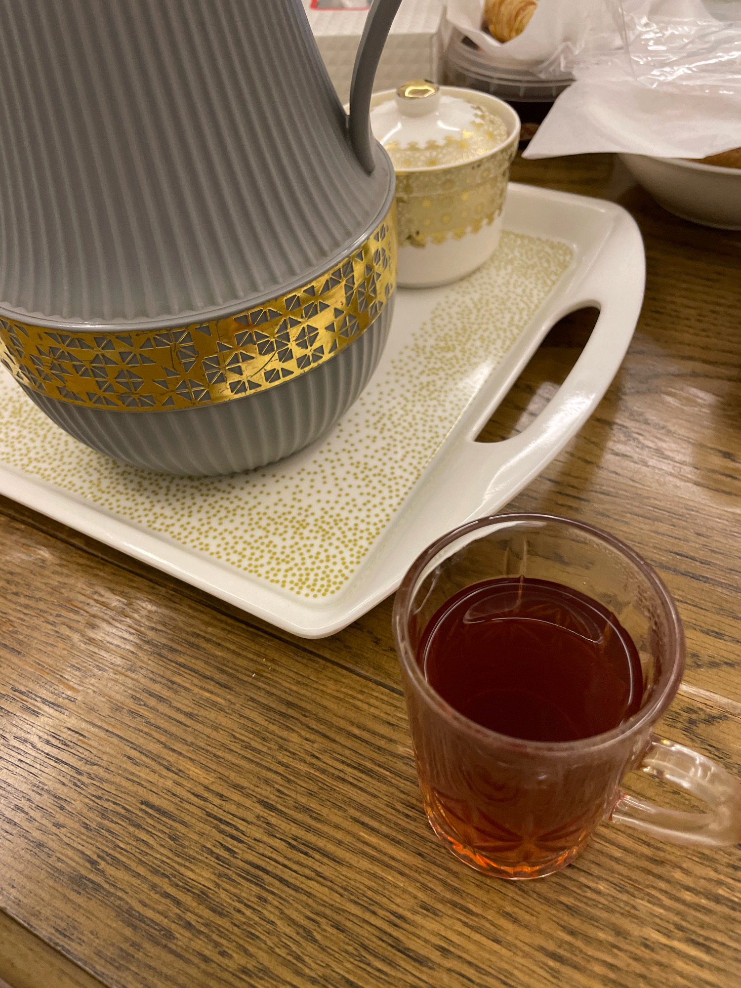 A close-up shot shows a glass cup of dark tea on a wooden table. To the left, a white tray with gold speckles holds a grey thermos with a gold patterned band and a small white and gold lidded dish. In the background, out of focus, are some pastries and white packaging. This scene is located in Umm Al Quwain, United Arab Emirates.