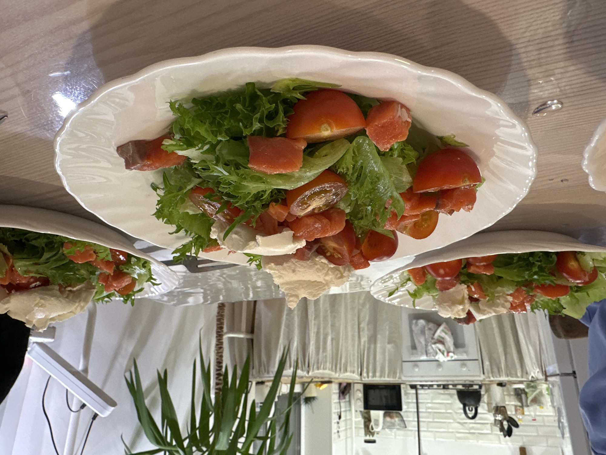 A high-angle, indoor shot depicts three white plates of salad. The salad contains green leafy lettuce, diced salmon, halved cherry tomatoes, and dollops of white cheese. The plates are arranged on a light-colored wooden surface. A green potted plant is visible in the foreground. The backdrop includes a kitchen area with white cabinets, a microwave, and a hanging kitchen scale. The location is Chaikovsky, Russia.