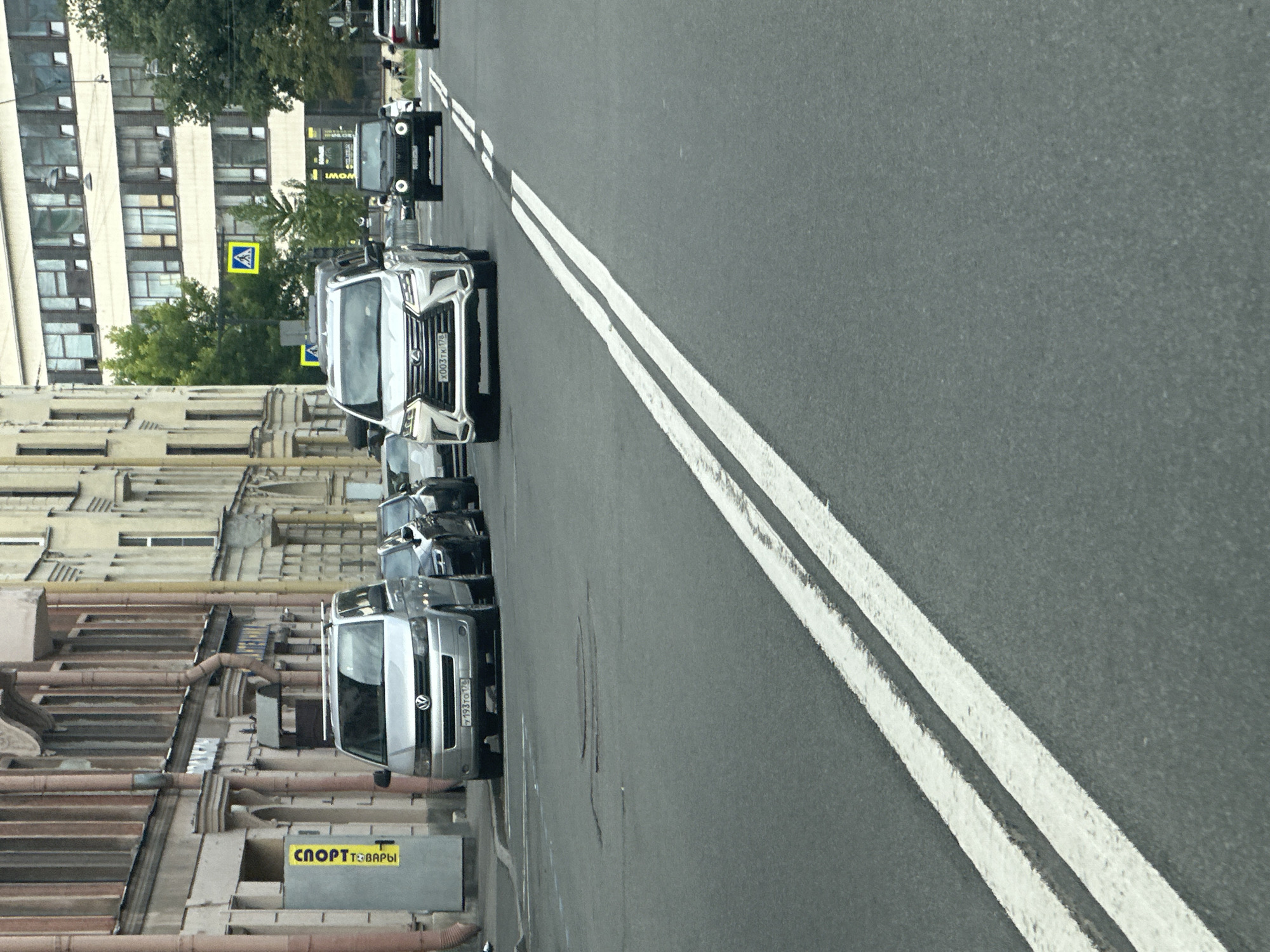 The image depicts a street scene in Saint Petersburg, Russia. In the foreground, a gray asphalt road is visible with double white lane markings. Several cars are present on the road. On the left side of the street, a silver Volkswagen minivan is parked, with a yellow sign reading "СПОРТ ТОВАРЫ" (Sports Goods) on a gray structure behind it. To the right of the minivan, a black Audi is parked, followed by another black vehicle. Further down the street, a white Lexus SUV is driving, followed by a black Jeep. In the background, buildings with multiple windows line the street. A pedestrian crossing sign is visible on the right side of the street. The sky is overcast, suggesting a typical Saint Petersburg day.