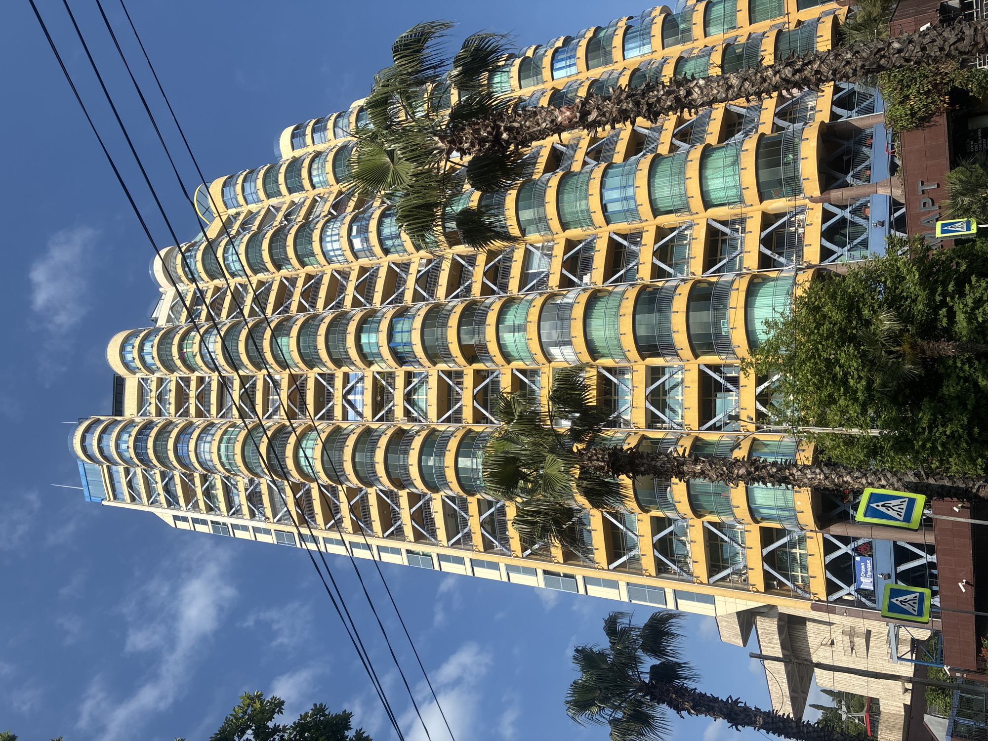 A tall, modern, yellow building with a curved facade and many rounded balconies is captured from a low angle, looking upwards. The balconies are lined with glass and appear to have light green curtains visible through them. The building's structure is accentuated by diagonal white supports. Two palm trees are in the foreground, one tall and slender with its fronds obscuring part of the building, and another shorter, thicker one on the right. Power lines are visible crisscrossing the bright blue sky, with a few wispy white clouds.
In the lower right portion of the image, there are two yellow and blue pedestrian crossing signs. Behind the building, a darker, brown-colored building is partially visible. The sky suggests a bright, sunny day. The overall scene evokes a warm climate, possibly a resort or coastal city. The building's unique architecture suggests a contemporary design.
The word "ОПТ" is visible on the brown building, possibly part of a larger sign.