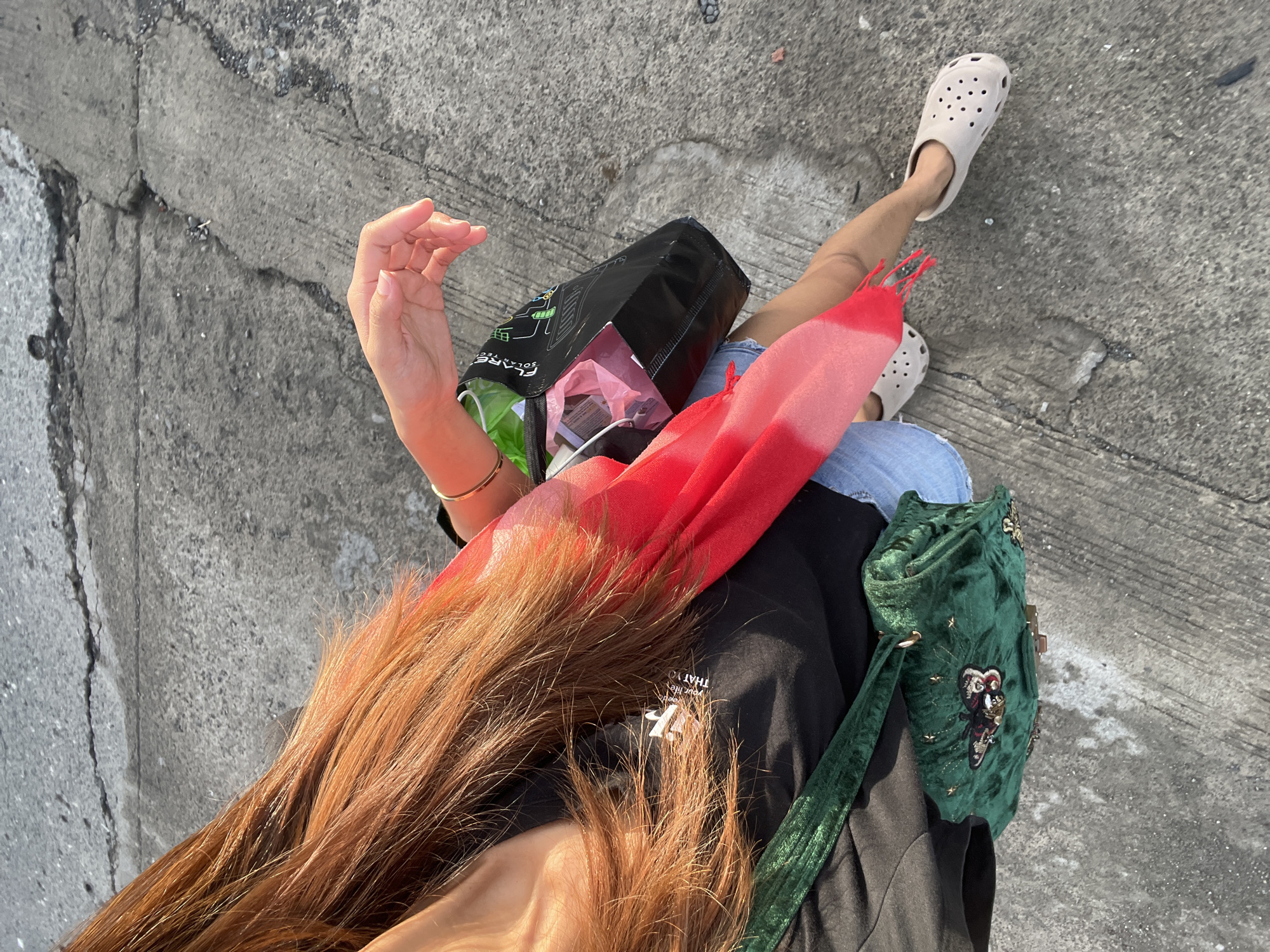 The image is a high-angle shot, seemingly taken by someone looking down at themselves. The person is standing on a cracked concrete surface, which appears to be a sidewalk or road.
A person's right hand is visible, fingers slightly curved. The person has auburn hair which falls over their shoulder. The person wears a bangle on their right wrist.
The person is wearing a black t-shirt, light blue jean shorts and beige crocs. They have a green velvet purse with gold accents on the right side of their body. There is also a red scarf and a black shopping bag. Inside the shopping bag there appears to be various objects. The shopping bag is printed with white symbols and text on the outside.
The setting is likely an urban area. It is not possible to conclusively confirm the location as Davao City, Philippines, based solely on the image.