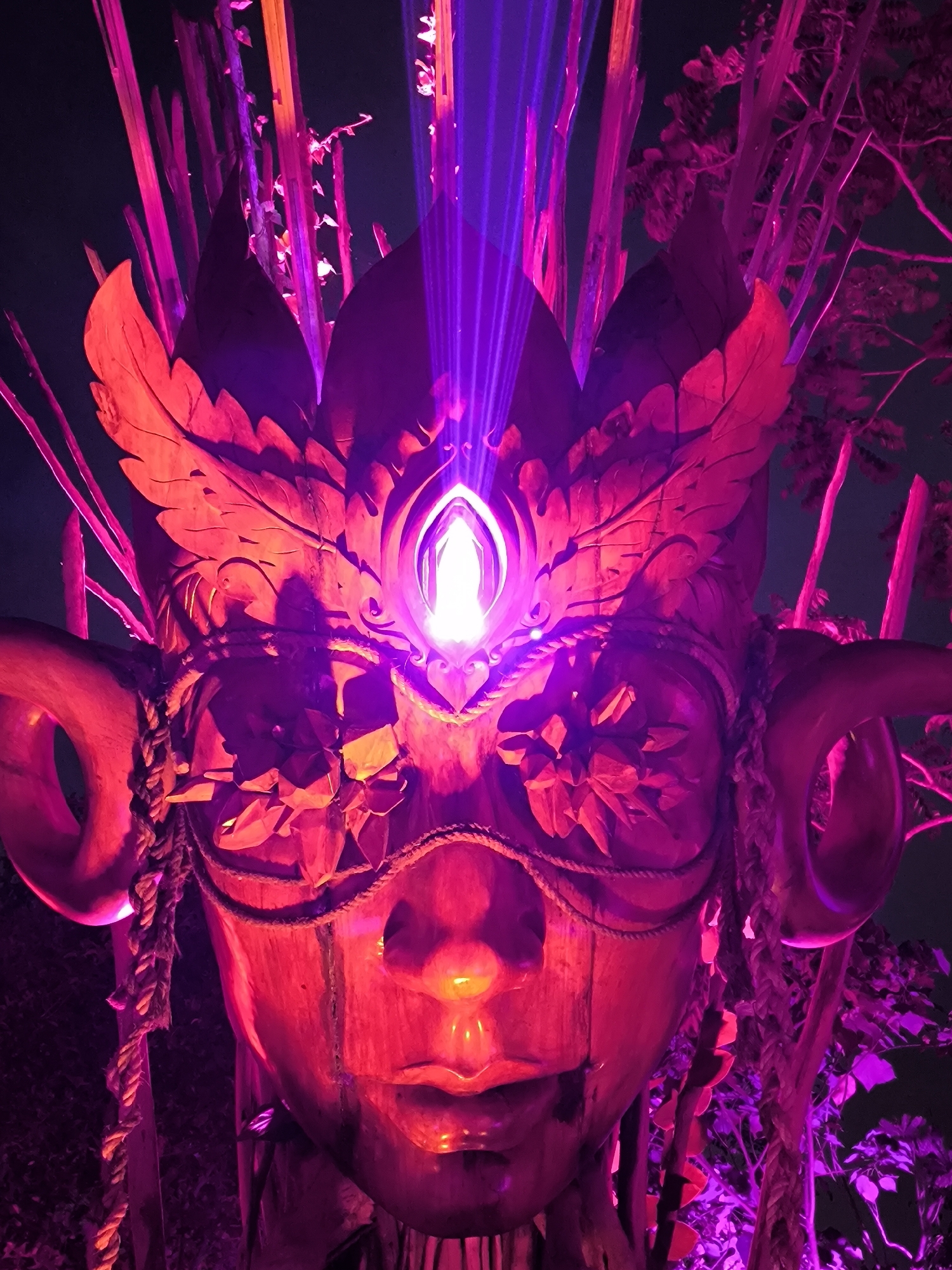The image depicts a large, ornate mask, likely a prop or installation, bathed in vibrant purple and pink light. The mask appears to be made of wood or a similar material and is sculpted to resemble a face with prominent horns. It is adorned with decorative elements, including what looks like carved leaves and a central, gem-like object at the forehead that is emitting a bright, luminous beam of light upwards. The mask is surrounded by dark, possibly night, surroundings, with vertical elements that could be trees or bamboo stalks, also illuminated by the colored lights. The overall atmosphere is mystical and somewhat theatrical. There is no indication of the city of Kediri, Indonesia, within the image itself.