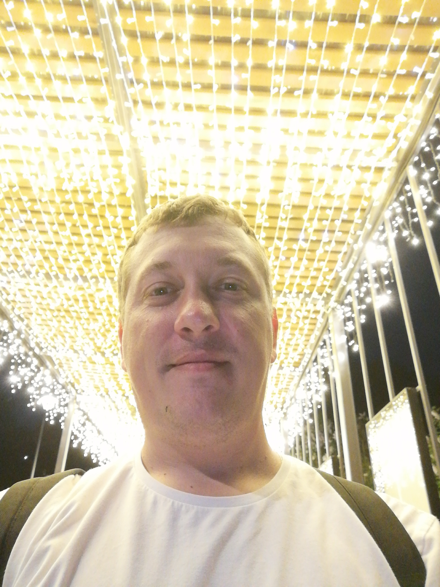 A medium shot captures a man with light skin, fair hair, and a white t-shirt, with a backpack obscuring the top of his shoulders. He is looking directly at the camera with a slight smile. Above him is a ceiling of densely packed, small, warm white lights arranged in a grid pattern. To the right of the man, vertical metal posts and more lights frame a dark backdrop. The location is identified as Unknown, Unknown.