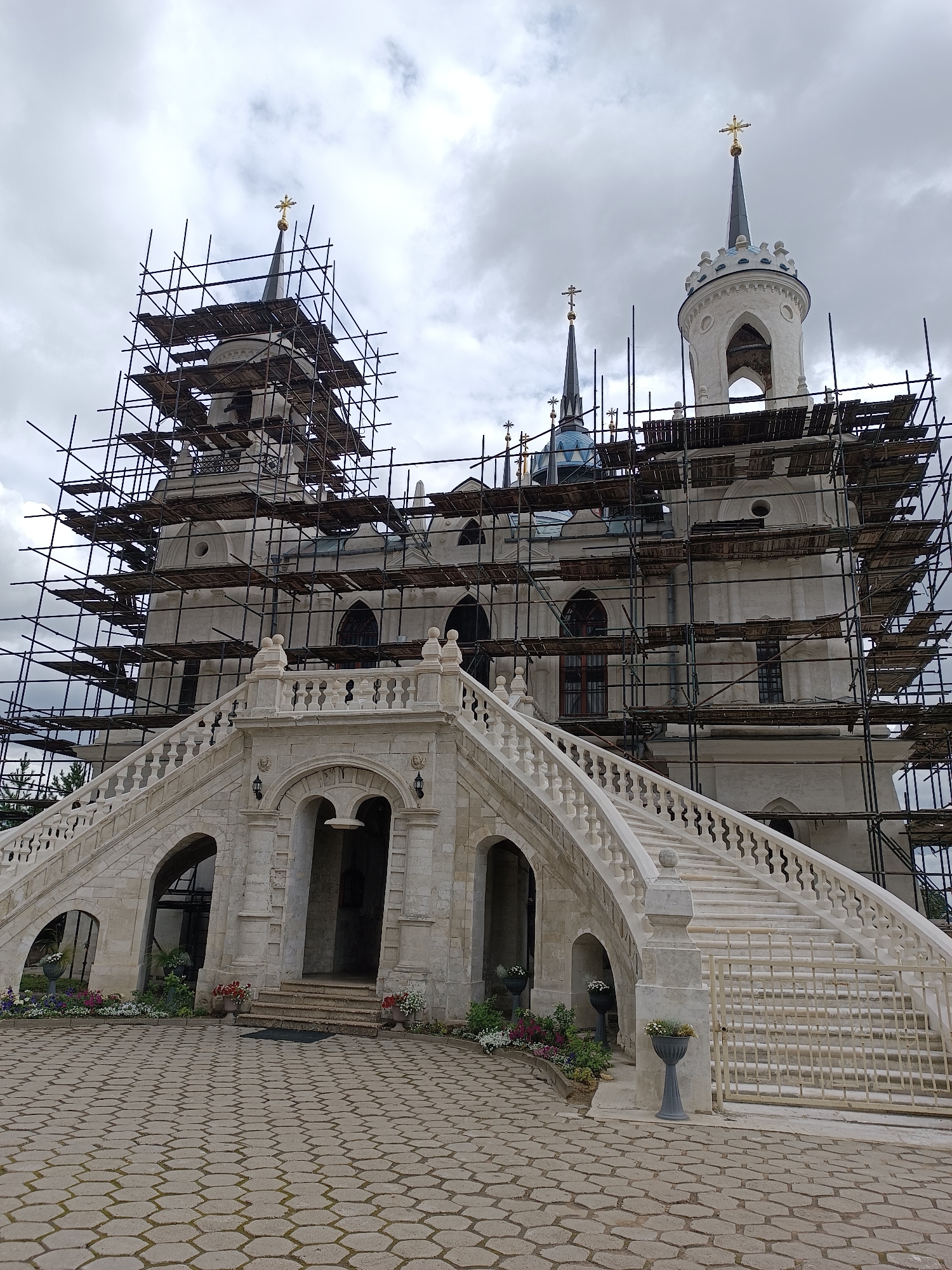 The image depicts the Church of the Vladimir Icon of the Mother of God in Bykovo, Russia, undergoing renovation, as evidenced by the extensive scaffolding surrounding its facade and towers. The white stone church features multiple spires, some topped with golden crosses. A grand, ornate white stone staircase leads up to the main entrance, which is arched and recessed. Planters with colorful flowers and greenery are arranged around the base of the staircase and the surrounding paved courtyard. The sky is overcast with clouds. No people are visible in the frame.