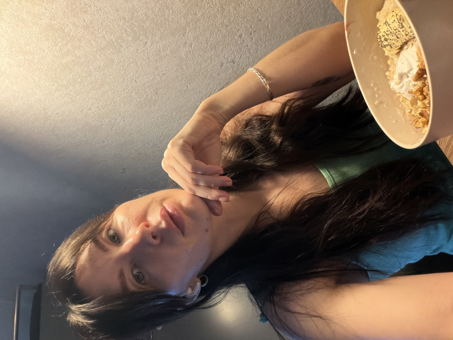 An indoor portrait depicts a female individual with long, dark hair, wearing a light green tank top. Small hoop earrings are visible on both ears, and a silver-colored braided bracelet is on her right wrist. Her chin rests on her left hand, which is raised to her face, with fingers slightly curled. She is looking directly towards the viewer. In the lower right foreground, a light-colored, oval bowl containing a meal, consisting of a base likely oatmeal or porridge, topped with walnuts, a dollop of white cream, a slice of banana, and chia seeds, is situated on a wooden surface. The background is a plain, textured grey wall. The specific location within Perm, Russia, cannot be determined from the visual information provided in the image.