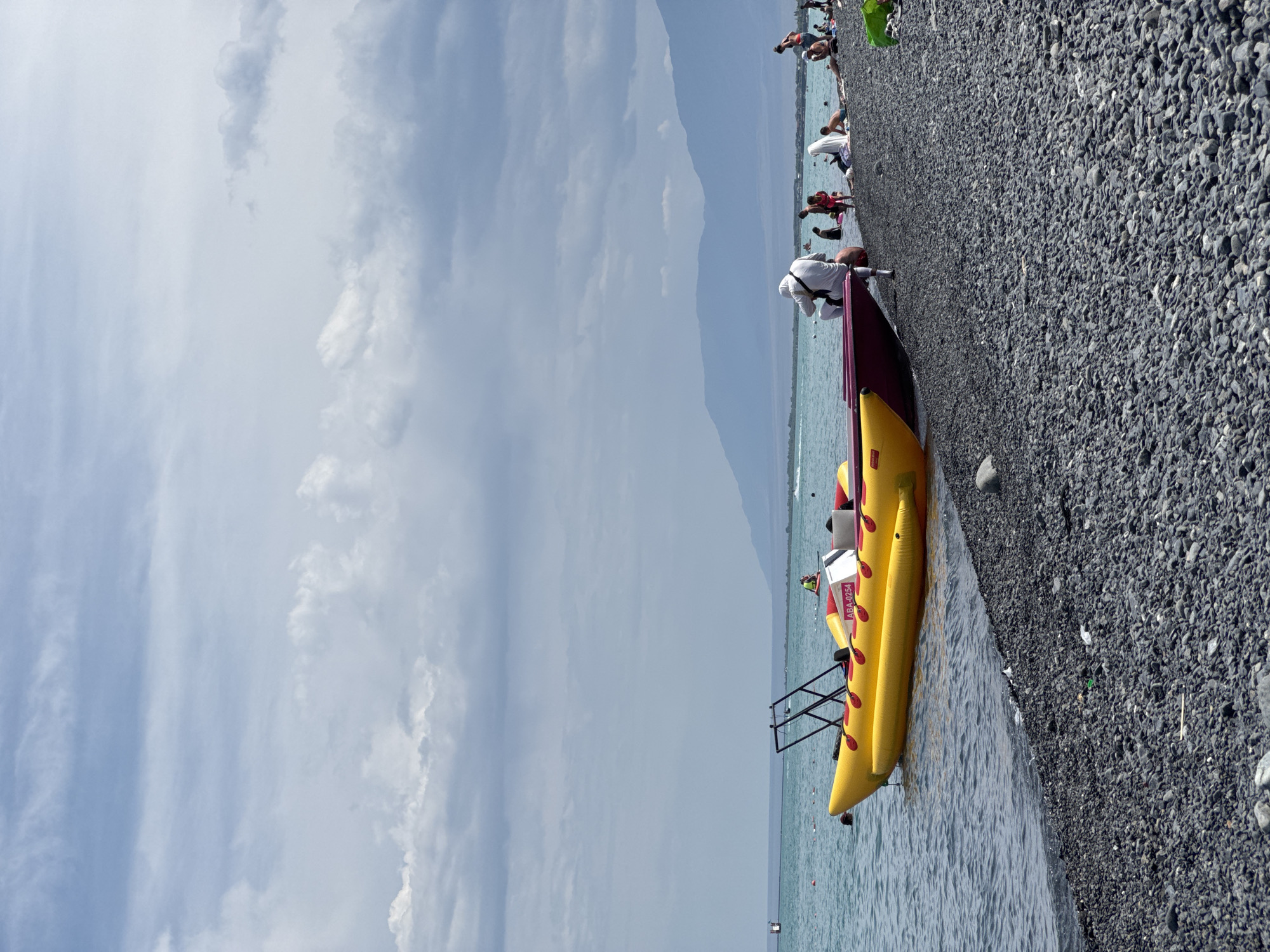 An eye-level, medium shot captures a pebble beach with a yellow and red inflatable boat docked near the shore. The boat has the designation "ABA-0254" on its side. The water is a clear turquoise, and in the distance, a range of hazy blue mountains stretches across the horizon under a cloudy sky. Several people are visible on the beach, some sitting and others standing. A few individuals are also in the water. The scene appears to be a recreational area, likely a beach resort or park, in the city of Ақши, Kazakhstan.