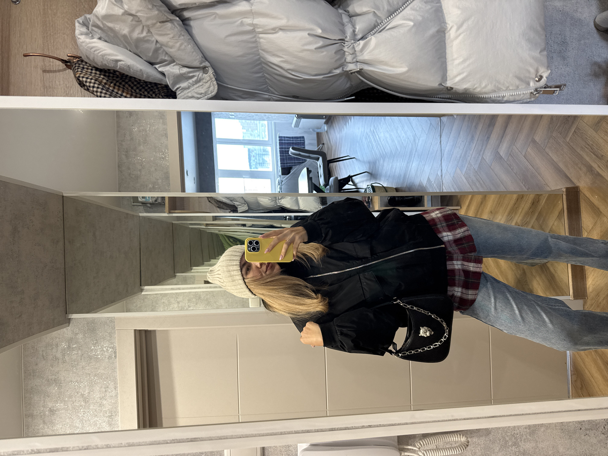A woman is taking a mirror selfie. The background is a modern interior, with gray walls and beige doors. The woman is wearing a white beanie, a black jacket, a red and black plaid shirt, and blue jeans. She is holding a yellow phone in her right hand, taking the picture. A small black purse hangs from her shoulder. Above, in the top of the mirror's reflection, a silver puffer jacket is hung up, and further in the background, a window shows a view into another room with a desk and chair. The flooring visible in the reflection is a light-colored wood with a herringbone pattern.