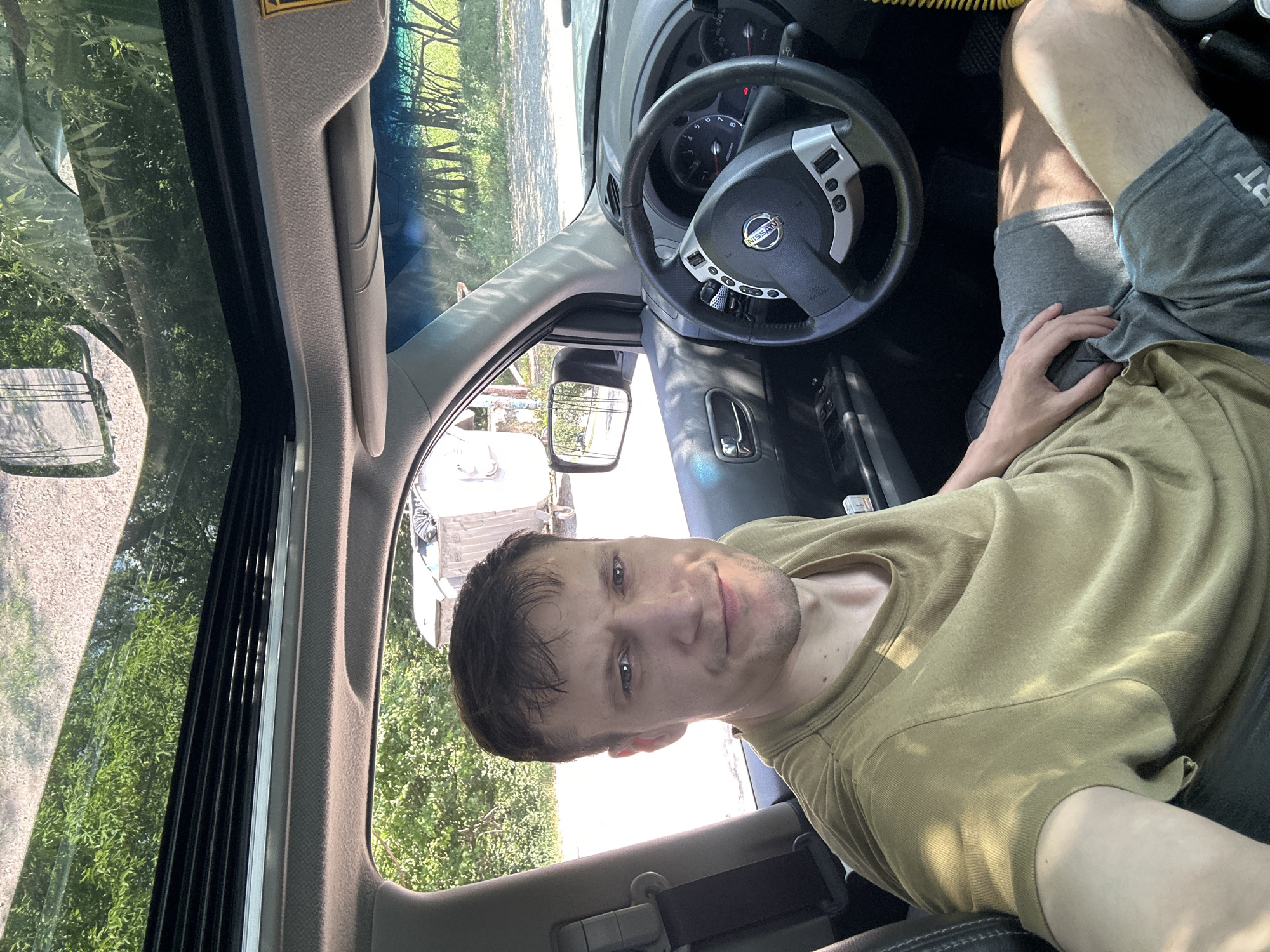 A medium shot captures a man sitting in the driver's seat of a Nissan vehicle, looking directly at the camera with a slight smile. He is wearing a khaki t-shirt and grey shorts. The car's interior is visible, including the steering wheel, dashboard, and a sunroof, which is open, revealing trees and a bright sky. Outside the car, a rural landscape with trees and some structures can be seen, suggesting a location on the outskirts of the city of Makiv, Ukraine. The lighting suggests it is daytime with sunlight streaming in.