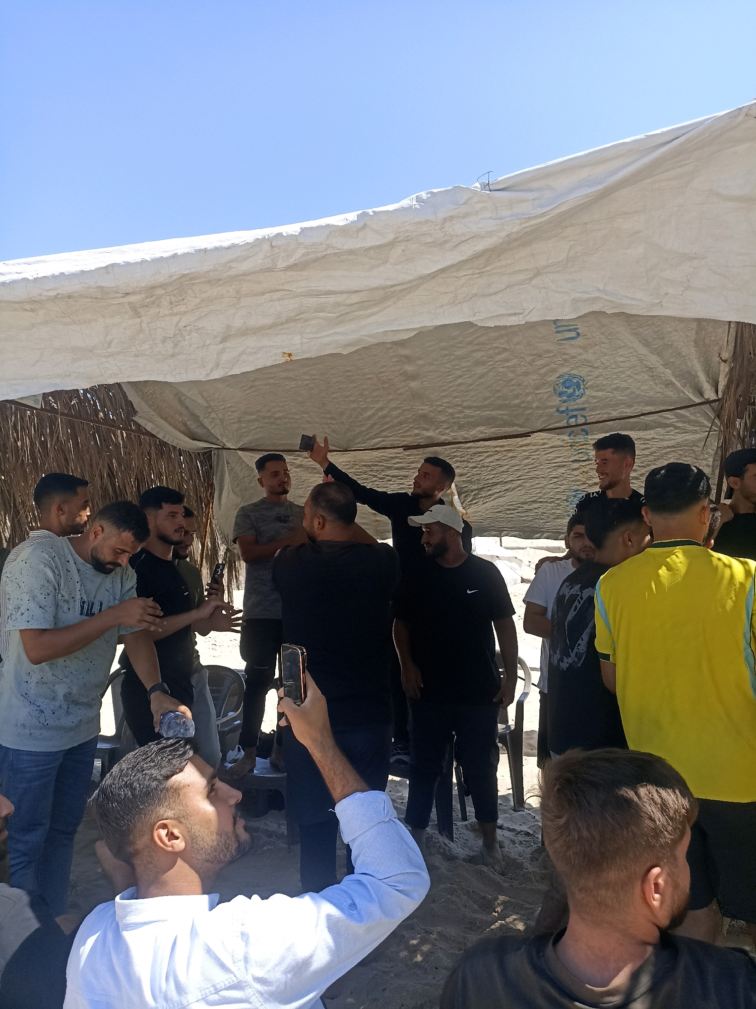 In Gaza City, Palestinian Territories, a group of men are gathered under a white tarp shelter. The shelter is made of a large piece of fabric stretched between poles, with palm fronds visible in the background. The ground is covered in sand. Several men are holding mobile phones, with one man in the foreground taking a selfie. Another man is holding up a bottle of water. The men are dressed in casual clothing, including t-shirts and jeans. The sky is clear and blue. The overall scene suggests a casual gathering or outdoor event.