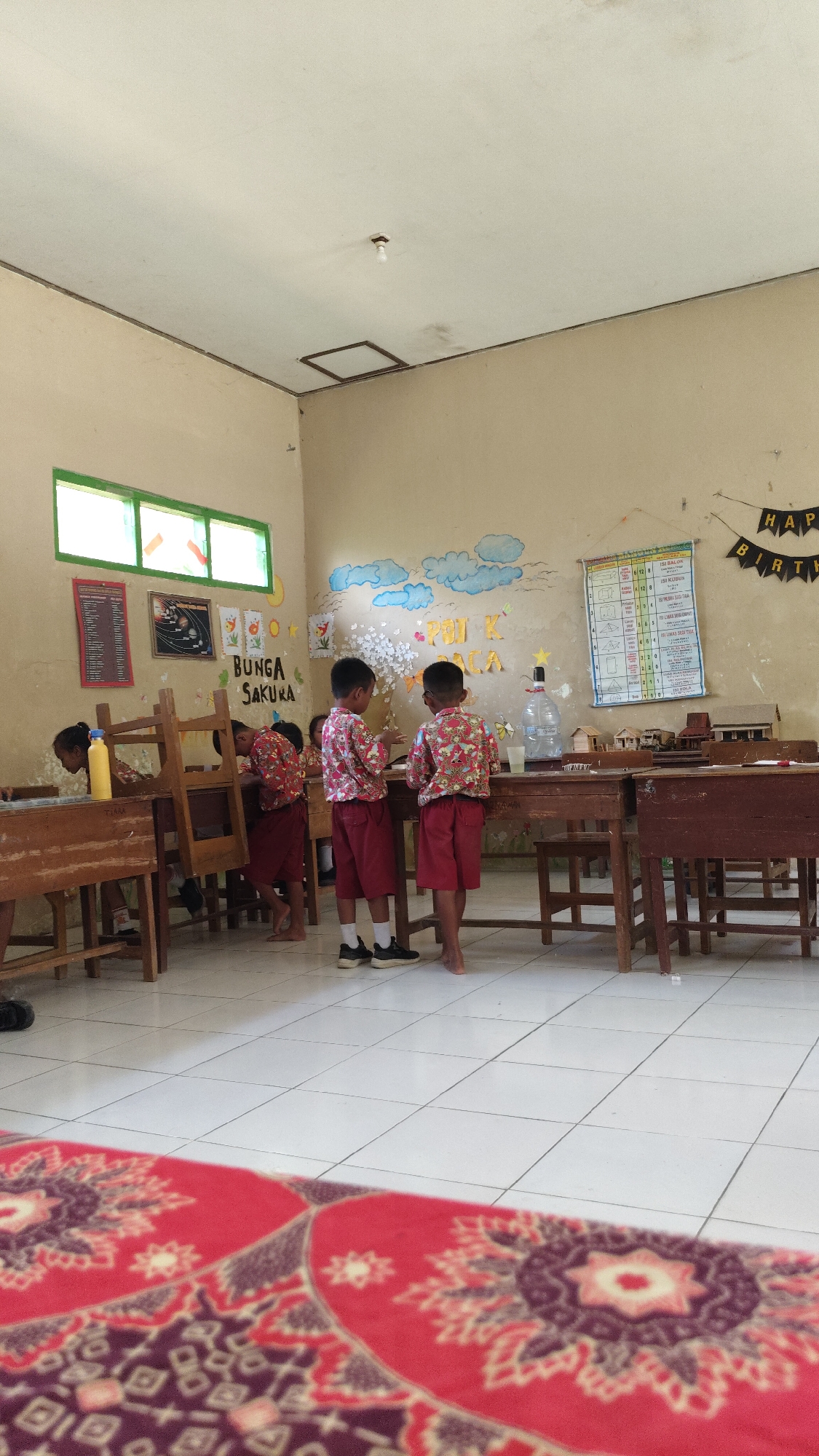 This image depicts an indoor scene, likely a classroom in Banjarsari, Indonesia. Two young boys, dressed in red shorts and patterned shirts, stand facing a table with a large water bottle and a few small models of buildings. Their backs are to the camera. In the background, other children are partially visible, with one appearing to be near a desk on the left side of the frame. The walls are a light tan color and decorated with children's artwork, including clouds and some text. A birthday banner with "HAPPY BIRTH" is visible on the upper right wall. There are multiple wooden desks and chairs arranged throughout the room. The floor is tiled in white squares. A red and purple patterned rug is in the foreground, partially obscuring the view of the floor. A window with green frames is visible on the left side of the image.