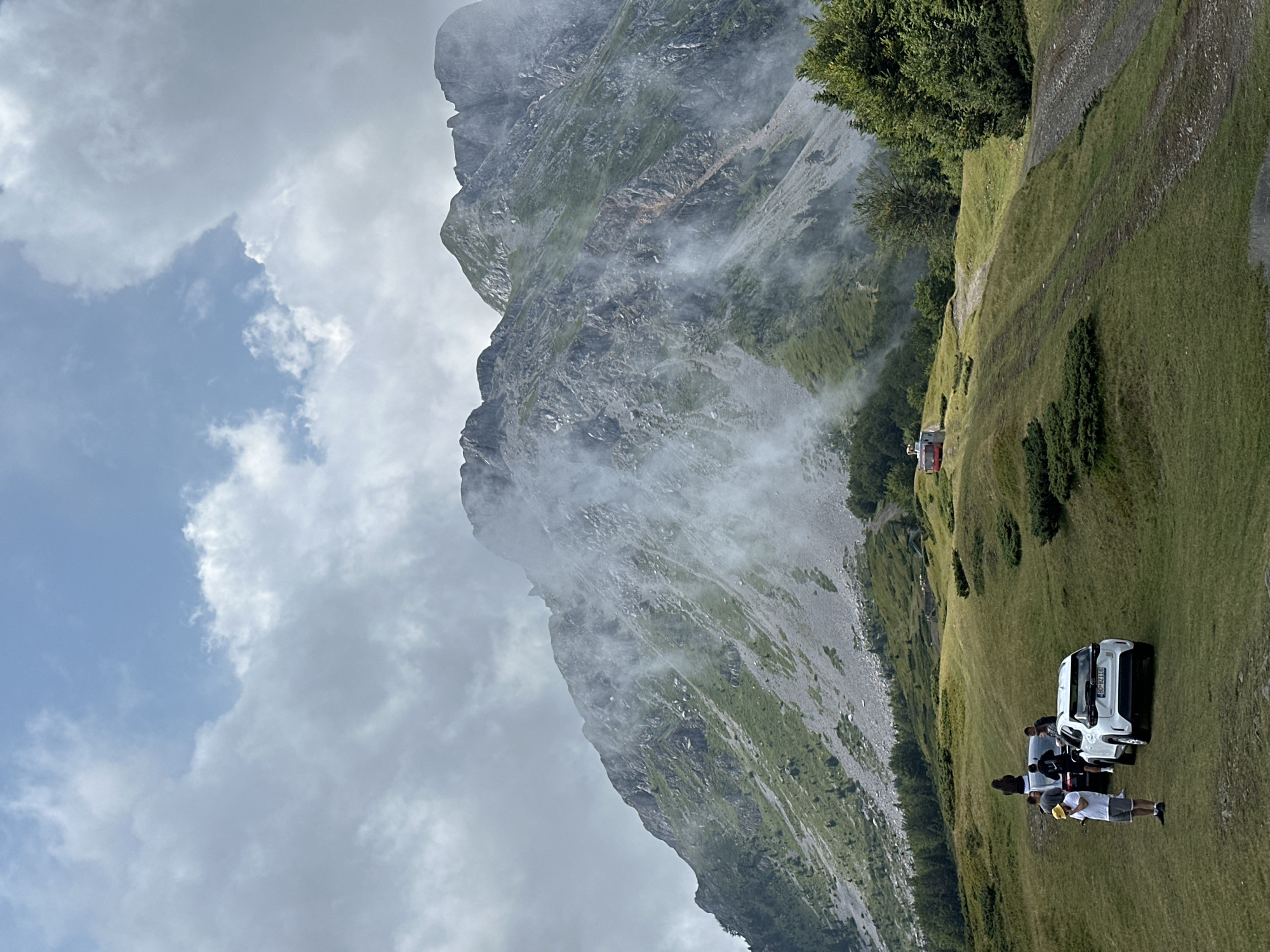 In the city of Unknown, Montenegro, a group of people are gathered in a mountainous, grassy terrain. The scene features several vehicles, including a white SUV and a dark sedan, parked on a grassy path. A man in a yellow hat and white t-shirt stands near the white SUV, while other individuals are positioned around the vehicles, some appearing to be interacting with each other or looking at their phones. In the background, a steep, rocky mountain slopes upward, partially shrouded in mist. Green trees and vegetation cover the lower parts of the mountain and the surrounding landscape. A small red structure, possibly a cable car station, is visible further up the hill. The sky above is a mix of blue and cloudy.