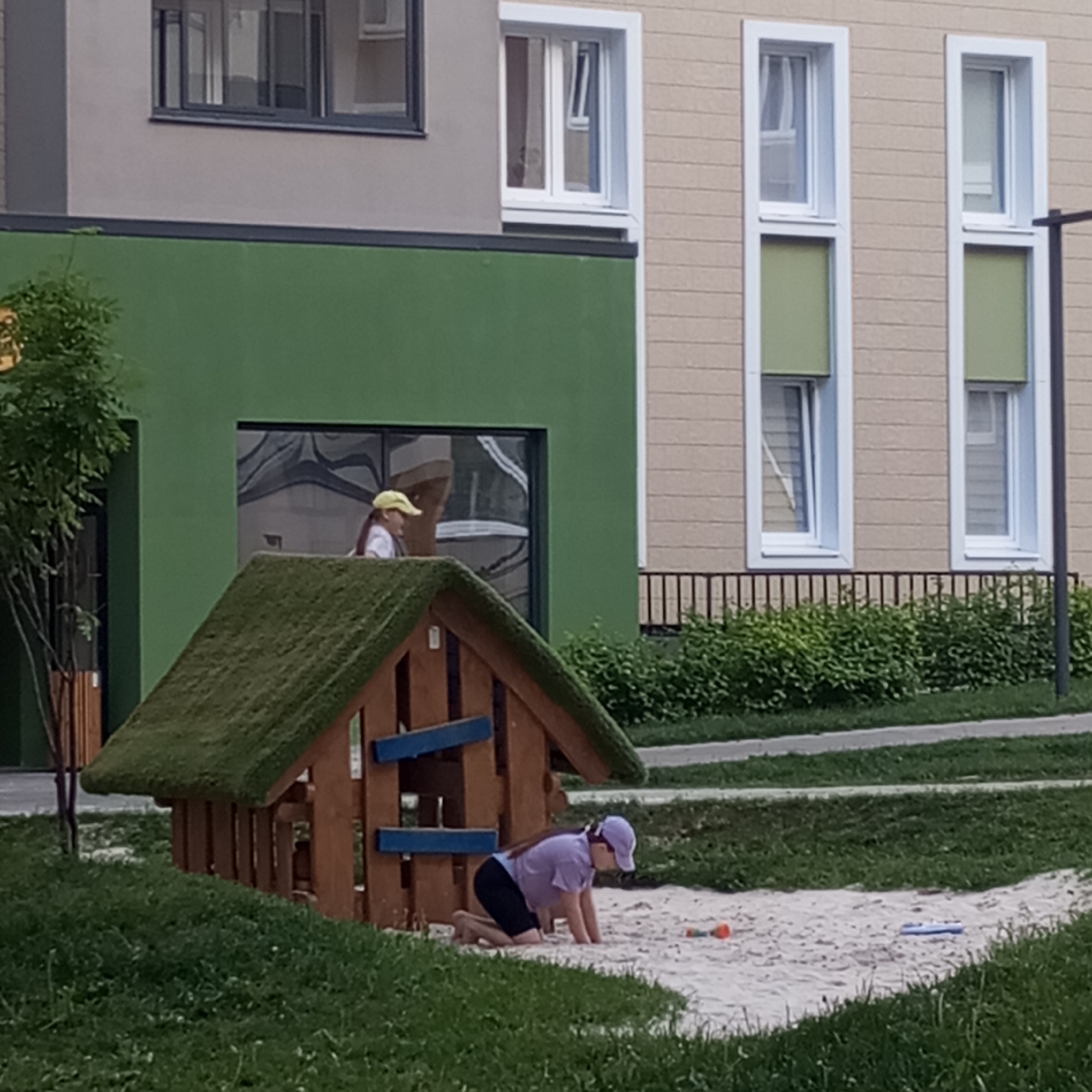 The image captures an outdoor scene in what appears to be a residential courtyard or playground. In the foreground, a young girl, wearing a purple cap, purple shirt, and dark shorts, kneels in a sandy area, engaged in play. Next to her is a wooden playhouse with a green, grass-like roof. Various small toys, including an orange object and a blue object, are visible in the sand.
In the mid-ground, another girl, wearing a yellow cap and a white shirt, is visible through a large window on the ground floor of a green-colored building. She appears to be standing or looking out, her reflection slightly visible in the glass. This green building has a dark grey upper section with windows.
In the background, there is a lighter beige or cream-colored building with multiple rectangular windows, some of which have light green blinds partially drawn. A black metal railing separates the buildings from the green lawn below. Lush green bushes and grass cover the ground, with a paved path or sidewalk running across the mid-ground. The overall lighting suggests it is daytime, possibly overcast, as there are no harsh shadows. No visible text is present in the image. The setting conveys a calm, everyday scene in a modern residential area in Pervomaysky, Russia.
