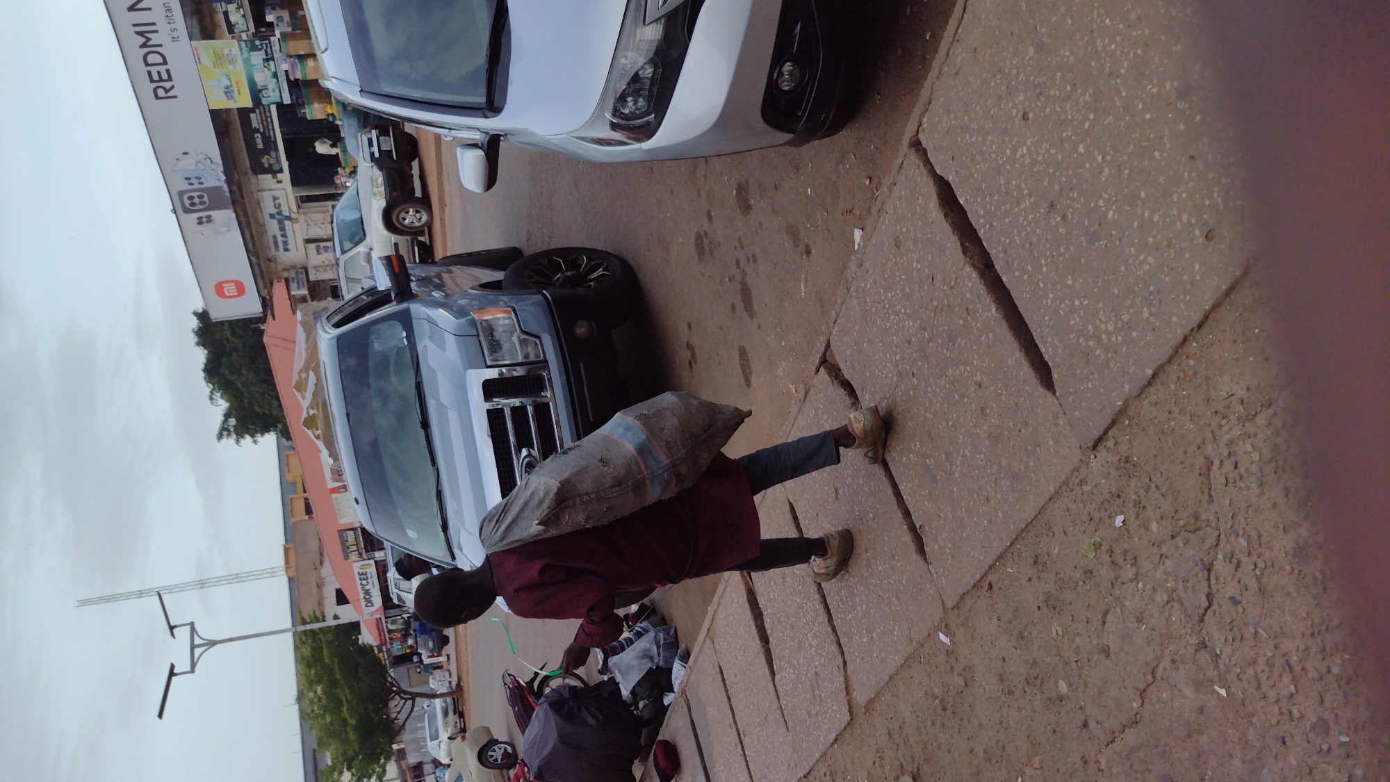 A young male is seen from the rear carrying a large, grey sack over his left shoulder as he walks along a concrete sidewalk. He is wearing a maroon long-sleeved shirt and dark trousers, with light-colored shoes. To his right, a grey Ford pickup truck is parked. To the right of the pickup truck, a silver sedan is also parked. In the background, there are several buildings with storefronts, including one with a "REDMI" advertisement. The scene appears to be a street in the city of Jos, Nigeria, with overcast skies.