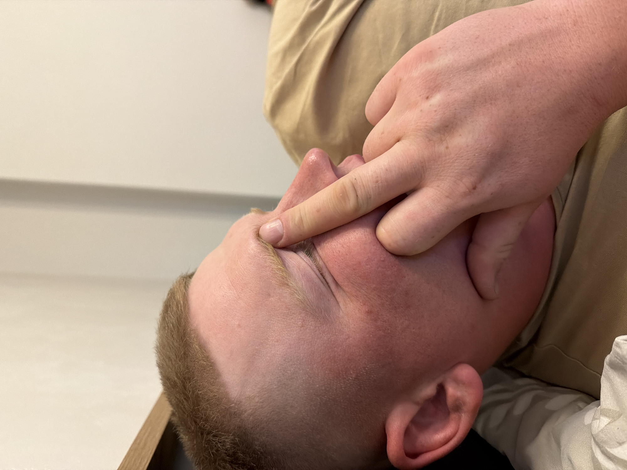 A close-up, tilted shot shows a person with light skin and short, blonde hair. Another person's hand is touching the first person's face near the eye area. The person is wearing a light tan shirt, and the background is a plain, light-colored wall. There is no indication of Naberezhnye Chelny, Russia, or any other location.