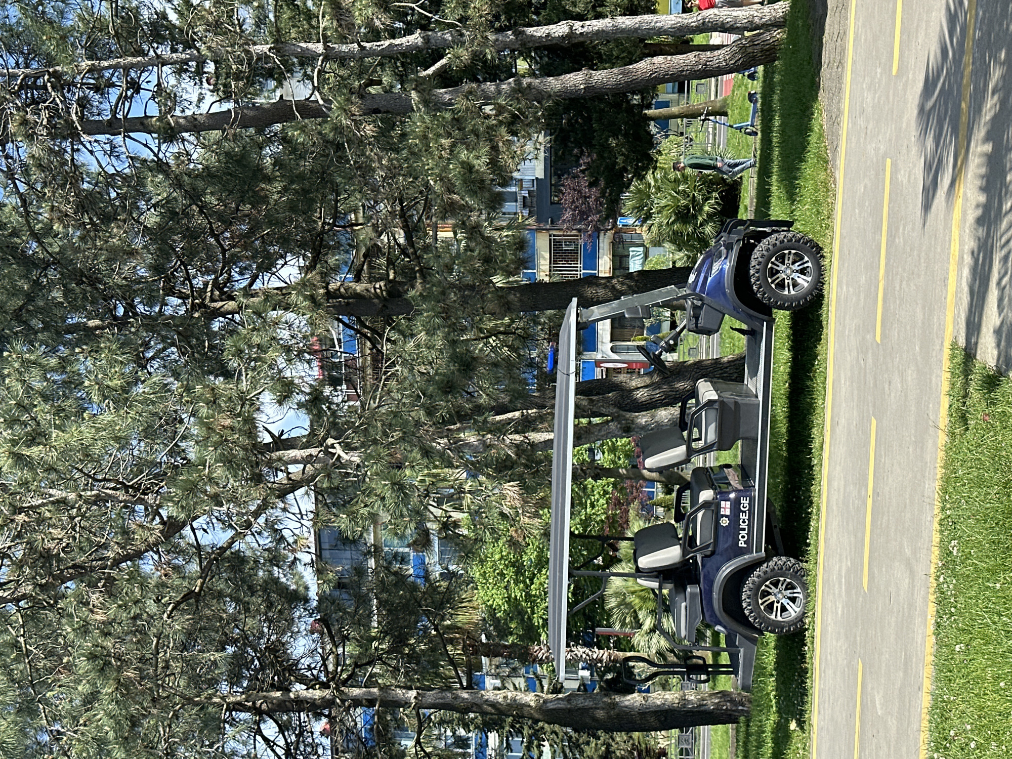 The image depicts a blue, four-seater golf cart with "POLICE.GE" markings parked on a paved path adjacent to a grassy area with trees. The cart is positioned on the right side of the frame, parallel to the path's edge, with its front facing towards the top of the image. The path has two yellow lines running along its length.
In the background, there are trees with dense foliage, partially obscuring a building with blue and white accents. Further in the background, a person can be seen standing near some exercise equipment. The scene suggests a park or recreational area. Given the "POLICE.GE" marking on the golf cart, the location is likely in Georgia, possibly in the city of Batumi.