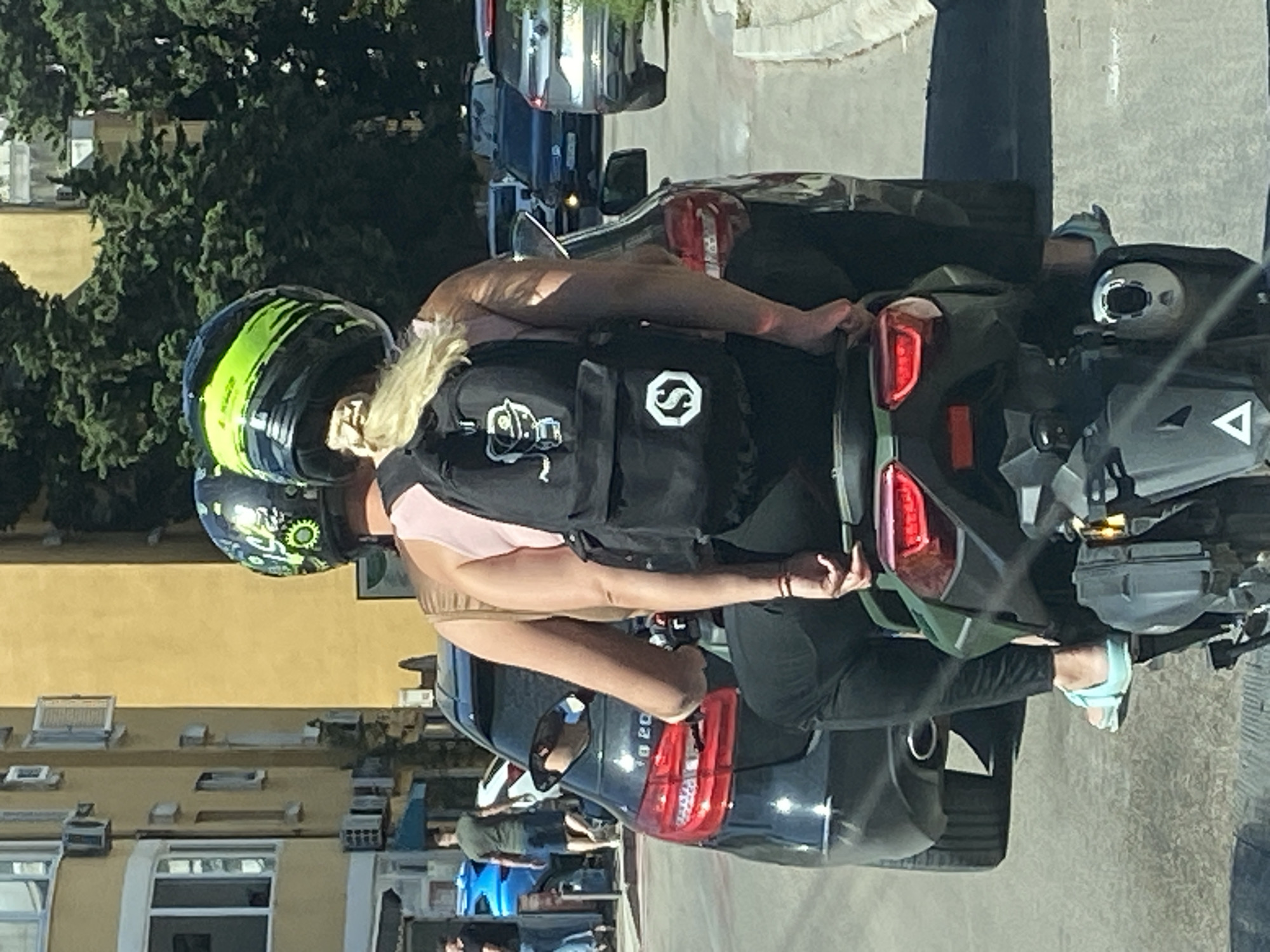 Two individuals are riding on a scooter in Budva, Montenegro. The person in the rear, who appears to be female, is wearing a pink tank top, black pants, and light blue sandals. She has blonde hair tied in a ponytail and is carrying a black backpack with a white graphic of a character and a circular logo. The person in front, wearing a black helmet with green accents, is also on the scooter. They are positioned behind a black Mercedes-Benz. In the background, there are buildings and trees, typical of an urban environment. The scene captures a moment of everyday life on a street in Budva.