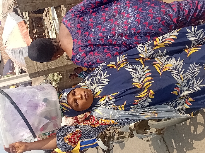 Two women are outdoors, one in a patterned blue and yellow hijab-like head covering, with a clear plastic container holding wrapped items next to her. She faces the camera, wearing colorful, patterned clothing. A second woman stands behind her, wearing a red and dark blue patterned dress. The background shows a building under construction and general outdoor clutter, along with another person partially visible further back.