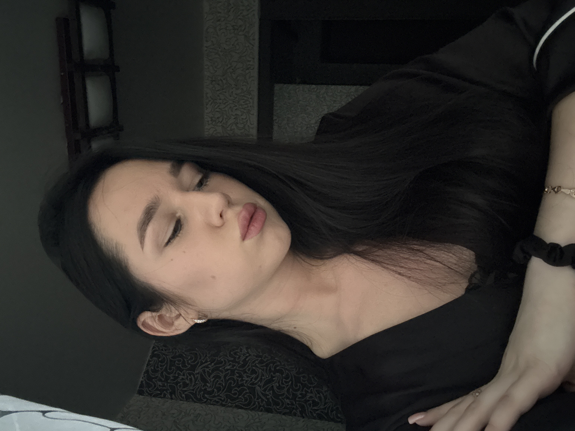 The image shows a close-up, waist-up, portrait of a young woman with long, dark hair, wearing a black satin robe. She is looking down and to her right, with her lips pursed. Her left arm is crossed in front of her chest, and she is wearing a small hoop earring in her visible ear. The background appears to be an indoor setting, with dark walls and what looks like a ceiling light fixture visible at the top. The lighting is dim and casts shadows. There are no identifiable landmarks or features that would indicate a specific location within Novosibirsk, Russia.