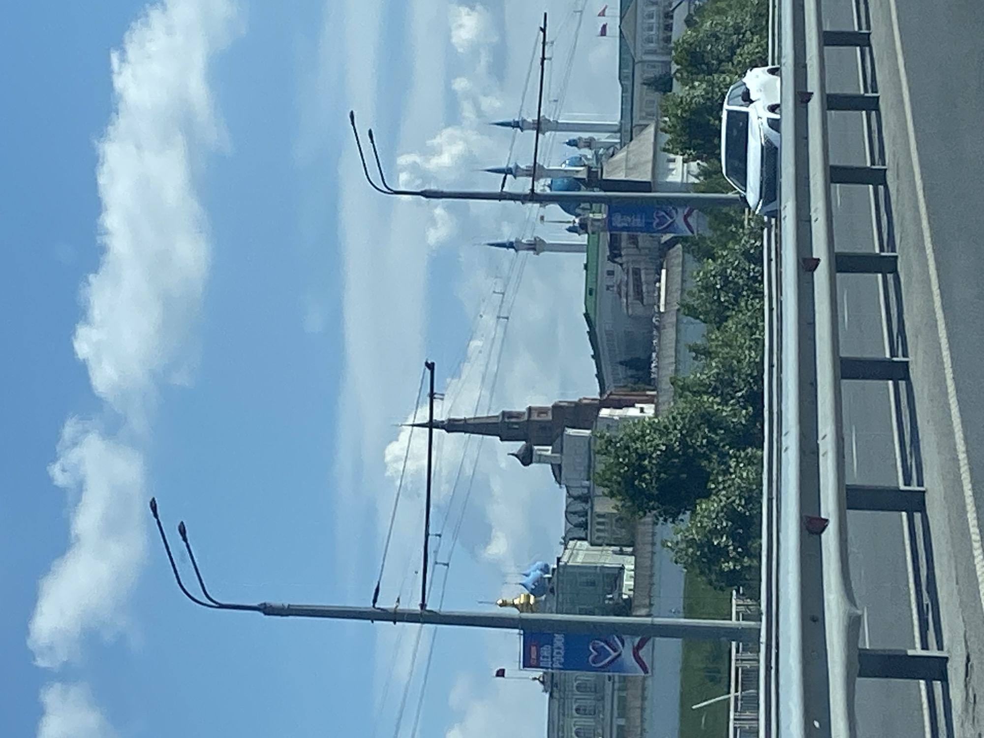 This is a medium-shot, eye-level, outdoor, daytime photograph of the city of Kazan, Russia, taken from an elevated position, likely a bridge or overpass. The foreground features a gray metal highway barrier with vertical supports casting shadows on the road below. A white SUV is partially visible on the road behind the barrier, its front end submerged as if it has driven off the road.
In the mid-ground, a dense line of green trees separates the road from a complex of historical buildings. Dominating the skyline are several prominent structures. To the left, a building with white facades and arched windows is visible, along with a tall, slender flagpole flying a red flag. A large blue banner with white text, including a heart symbol, is draped on the left side of this building. Adjacent to it are buildings with blue-domed roofs.
Towards the center and right of the image, a tall, multi-tiered reddish-brown tower with a pointed spire rises above the other structures. Further to the right, several white minarets with slender spires are visible, along with blue-domed buildings. These architectural elements suggest a blend of religious and civic architecture, characteristic of Kazan. Two smaller flags, red and white, are seen on a building to the far right.
The background is a bright blue sky filled with scattered white clouds. Two tall, modern streetlights with dual lamps are prominently positioned in the shot, one to the left and another towards the right, indicating the presence of modern infrastructure within the city. Power lines are strung between poles, crossing the scene horizontally. The overall impression is a view of Kazan's skyline, showcasing its historical landmarks against a backdrop of modern urban elements and a clear sky.