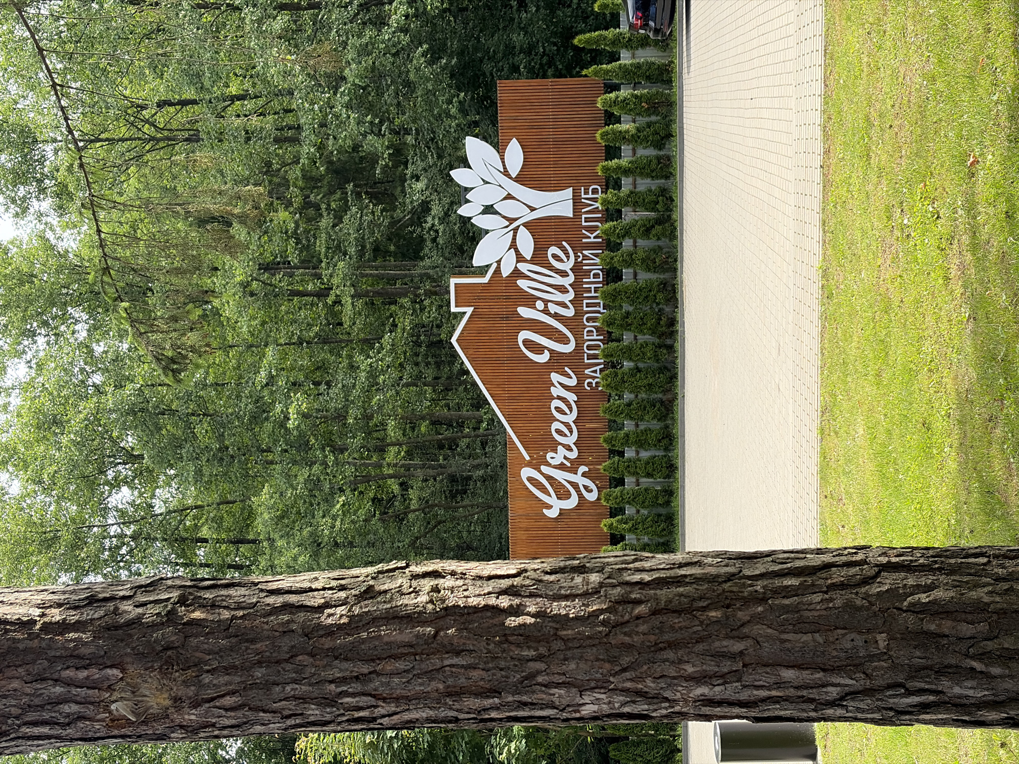 A wide shot captures a wooden sign for "Green Ville" in a rural Belarusian setting, likely within the Zhdanovichi rural council. The sign, featuring stylized white lettering and a house emblem with a tree, is mounted on a wooden plank background. The text "ЗАГОРОДНЫЙ КЛУБ" (which translates to "Country Club" in Russian) is written vertically along the right side of the sign.
The sign is surrounded by manicured green bushes, forming a neat border. To the right, a paved walkway runs alongside the bushes, and beyond that, a neatly mowed green lawn. The background is dominated by a dense forest of tall, leafy trees, suggesting a natural and tranquil environment. A partial view of a dark-colored car is visible on the far left, suggesting that this is an entrance to a property or establishment.
In the foreground, a large, rough-barked tree trunk spans the width of the image, creating a natural frame and adding a rustic element to the scene. The lighting indicates it's daytime, with bright sunshine casting shadows, and the lush greenery suggests pleasant weather. The overall impression is one of a well-maintained, upscale country club or residential area aiming for a blend of nature and comfort.