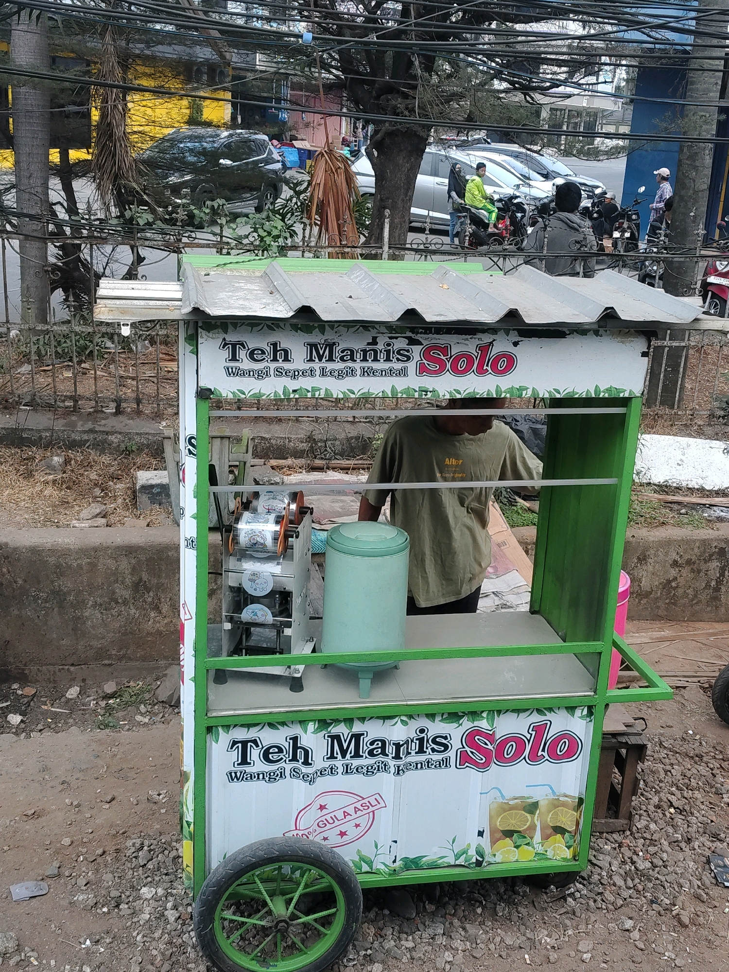 The image captures a street scene in Tangerang, Indonesia, focusing on a mobile beverage cart selling "Teh Manis Solo." The cart, adorned with green and white graphics, features the branding "Teh Manis Solo" and a tagline that reads "Wangi Sepet Legit Kental." Inside the cart, a person wearing a light green t-shirt is visible, possibly preparing drinks. Equipment for sealing cups is mounted on the cart, along with a large, light blue container. The cart is situated on a rocky, dirt surface next to a concrete barrier. In the background, a busy street can be observed with various vehicles, including cars and motorcycles, suggesting an urban environment. Power lines crisscross the sky, and trees with dry foliage line the area. The overall impression is that of a typical street-side vendor in a bustling Indonesian city.