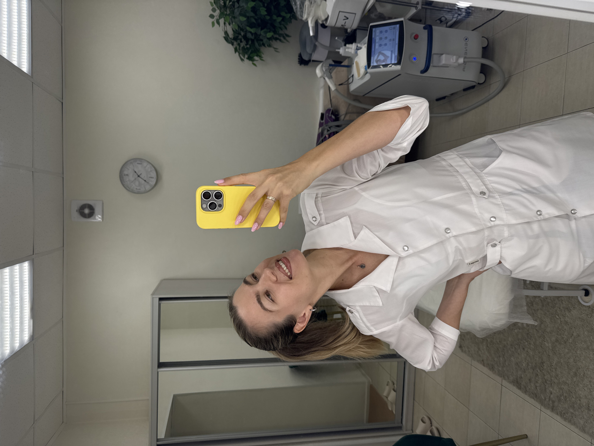 In an indoor setting, a Caucasian woman with blonde hair pulled back in a ponytail, wearing a white lab coat and a necklace, poses for a selfie taken with a yellow iPhone. She is smiling and looking at her phone. Her fingernails are painted pink. The background shows a room with a large mirror closet on the left, a clock on the wall, and what appears to be medical or aesthetic equipment on the right, including a white machine with a blue accent and a screen displaying various icons. A white examination table covered with plastic sheeting is visible behind the woman. The floor is tiled, and a grey rug is placed in front of the examination table. A green chair is partially visible on the far left. The lighting suggests an office or clinic environment.