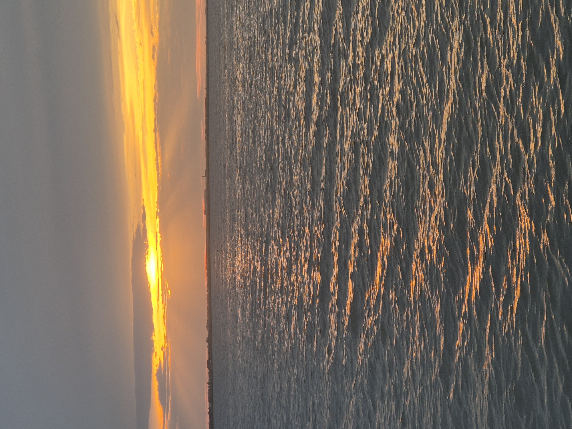 This image depicts a serene natural scene at sunset, possibly in Kokshetau, Kazakhstan. The horizon is divided into two distinct parts: the sky and the water. The left side of the image captures the sky, dominated by dramatic clouds bathed in the warm, vibrant hues of the setting sun. Streaks of bright orange and yellow cut through the muted grey of the upper sky, indicating the sun is low on the horizon. The right side of the image focuses on the water, which appears to be a large body like a lake or sea. The surface of the water is rippled, with the setting sun casting elongated, shimmering reflections that create a textured, almost abstract pattern of light and shadow. The overall mood is tranquil and contemplative, suggesting a moment of natural beauty. The time of day is clearly sunset, characterized by the intense colors in the sky and the long reflections on the water. No people, specific objects, or visible text are discernible in the image, allowing the focus to remain on the expansive natural landscape.