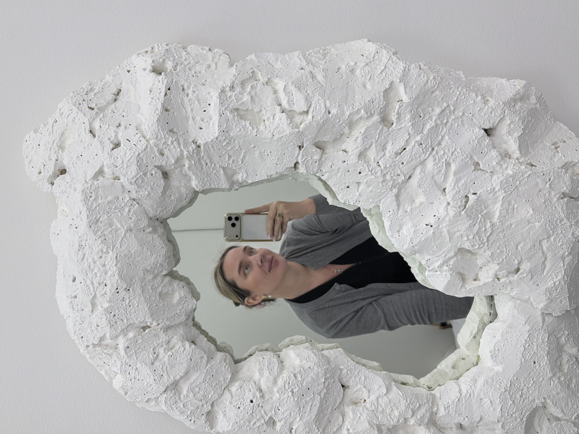 A woman is taking a selfie in a mirror. The mirror has a textured, white frame. The woman is wearing a gray cardigan and a black top. She is holding a cell phone in her right hand. The background is a plain, white wall. It is not possible to determine the location of the photo, including whether or not it is in Moscow, Russia.