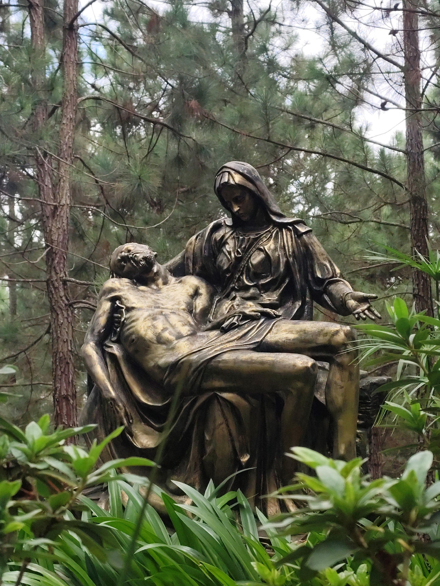 The image depicts a bronze sculpture of Mary holding the body of Jesus after the crucifixion, commonly known as the Pietà. The sculpture is situated outdoors in a wooded area with pine trees and lush green foliage in the foreground and background. Mary, with her head bowed and draped in robes, cradles Jesus, whose body shows signs of his suffering, including wounds. The sculpture appears weathered, suggesting it has been exposed to the elements. This particular Pietà sculpture is located at the Lạc Dương Church in Lạc Dương district, Lâm Đồng province, Vietnam, a popular pilgrimage site.
