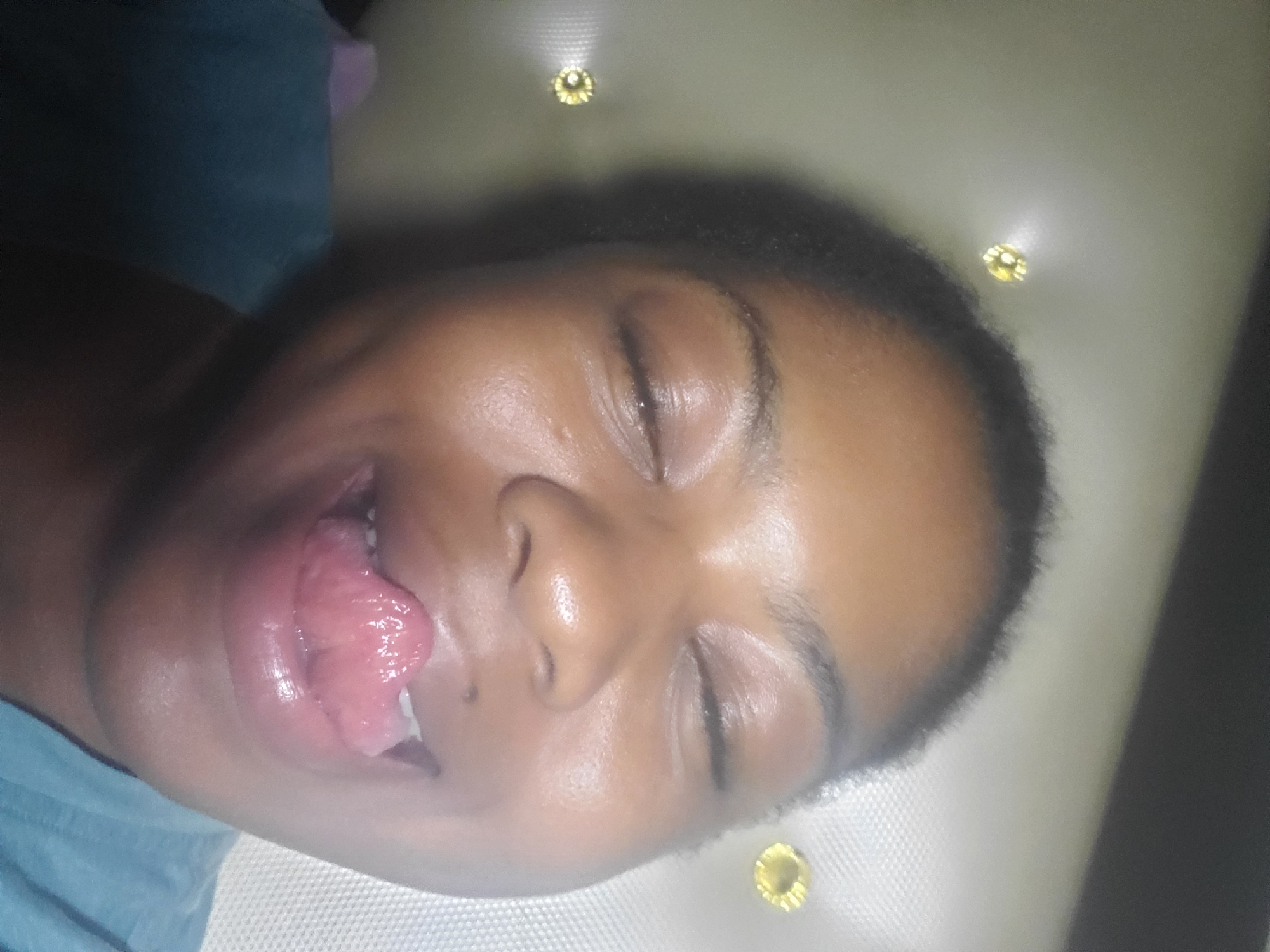 A close-up, eye-level indoor photograph taken in the city of Kakau, Nigeria, shows an individual with dark skin, eyes closed, and a wide smile. The person's mouth is open, with a pink tongue protruding and curled upwards. Visible are short, dark hair around the forehead and temples, and a portion of a blue-green garment on the right shoulder. The background features a light-colored, possibly upholstered surface, such as a headboard, with at least three circular, gold-colored decorative studs visible: two on the upper left and one on the upper right. The lighting is soft and even.