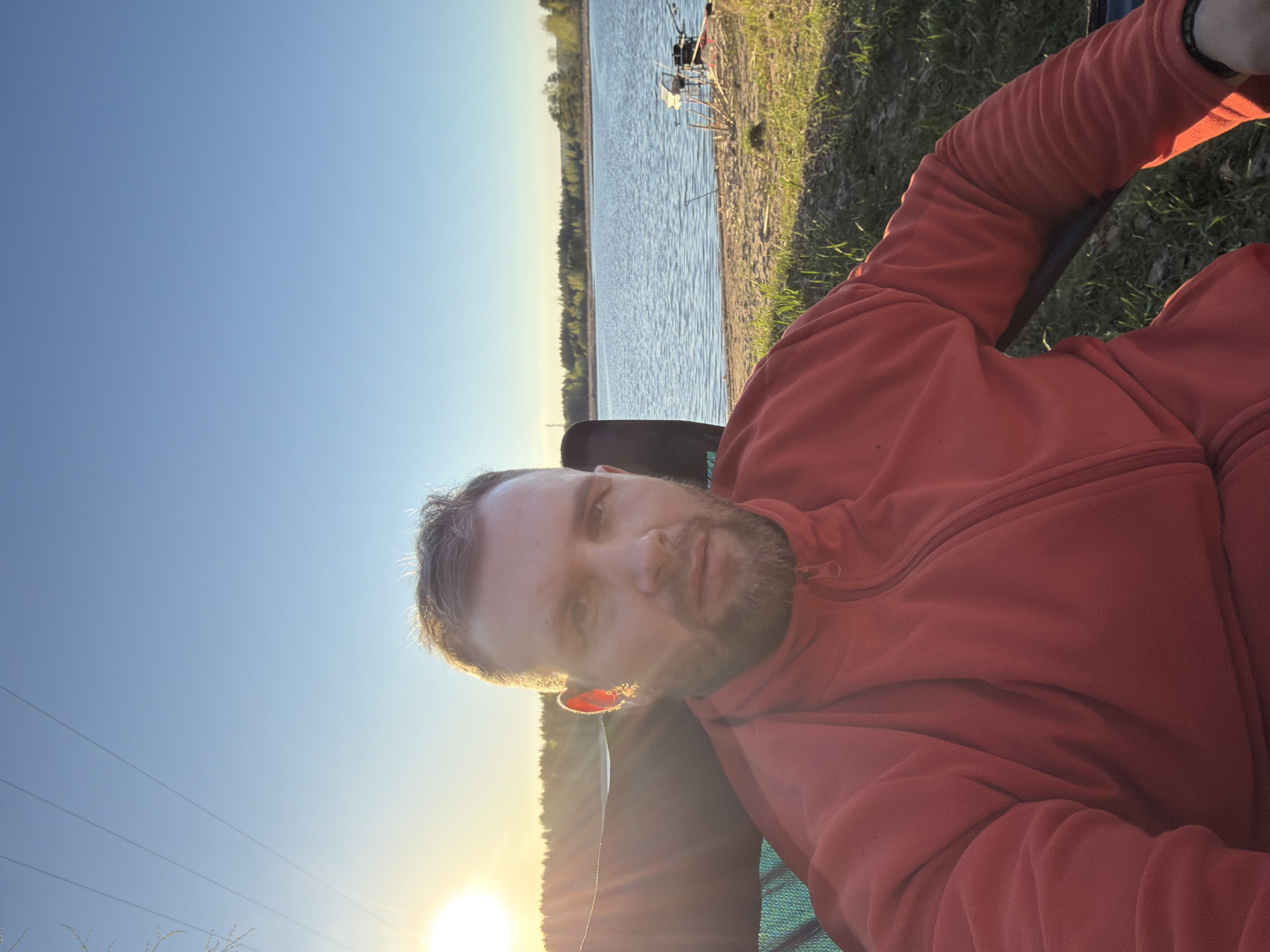 A man wearing a red fleece jacket is seated in a chair outdoors, facing the camera. The background shows a body of water, likely a lake or river, with trees lining the opposite shore. The sky is blue with a setting sun visible in the lower left corner of the frame. Fishing rods are set up near the water's edge. The location is near the water in Chaykovsky, Russia.