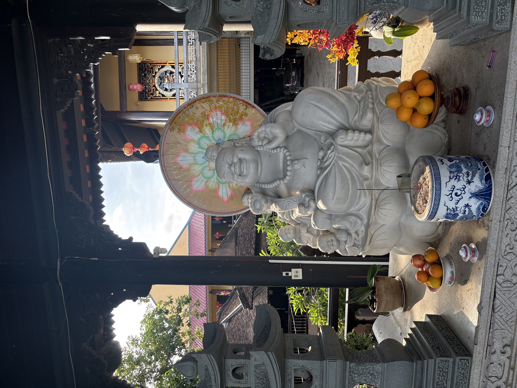 A stone statue of a laughing Buddha is the central subject of this outdoor scene. The Buddha statue is large and white, seated on a lotus base. Behind the Buddha, a circular, fan-like object with colorful patterns is visible. Several smaller, smiling stone figures are gathered around the Buddha's lap. In front of the statue, on a stone surface, is a small bowl containing several oranges and an apple, as well as a small tea set. A blue and white ceramic incense holder with lit incense sticks is also present. Two stone pagoda-like structures flank the Buddha statue. The background shows elements of a temple or pagoda, including tiled roofs, decorative wooden carvings, and colorful lanterns. A black car is visible in the distance. The overall environment appears to be a temple courtyard.