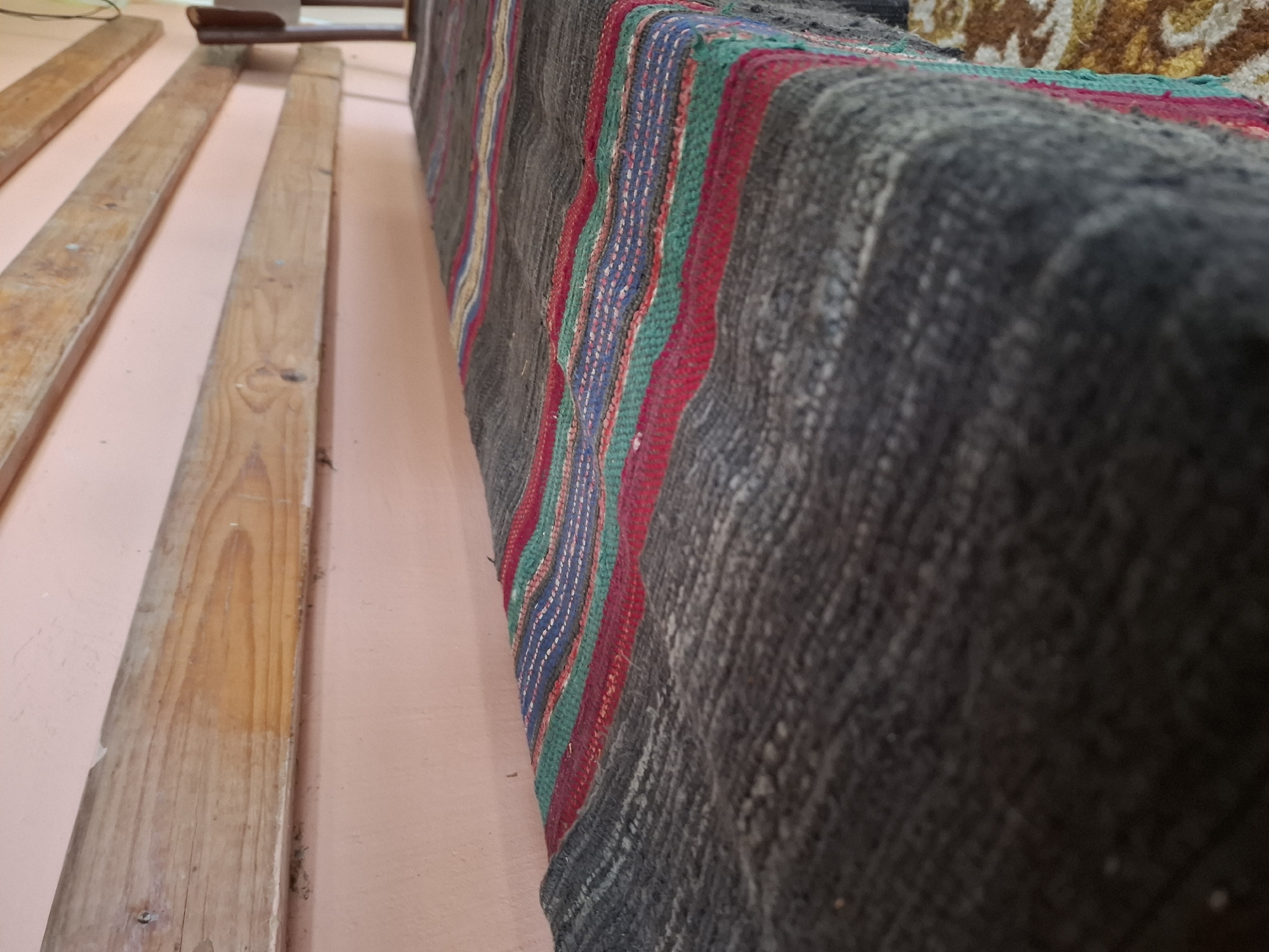 The image captures a close-up view of a woven blanket with stripes of red, green, and blue over a dark grey base. The blanket is laid out on a surface that appears to be a bench or a low table, partially visible in the foreground and to the right. The background consists of a pale pink wall with horizontal wooden planks mounted on it. The lighting suggests an indoor setting, possibly during the day, with a soft, diffused light source. The composition is focused on the texture and pattern of the blanket, with the background elements slightly out of focus. The image does not contain any information about the location in Malko Asenovo, Bulgaria.