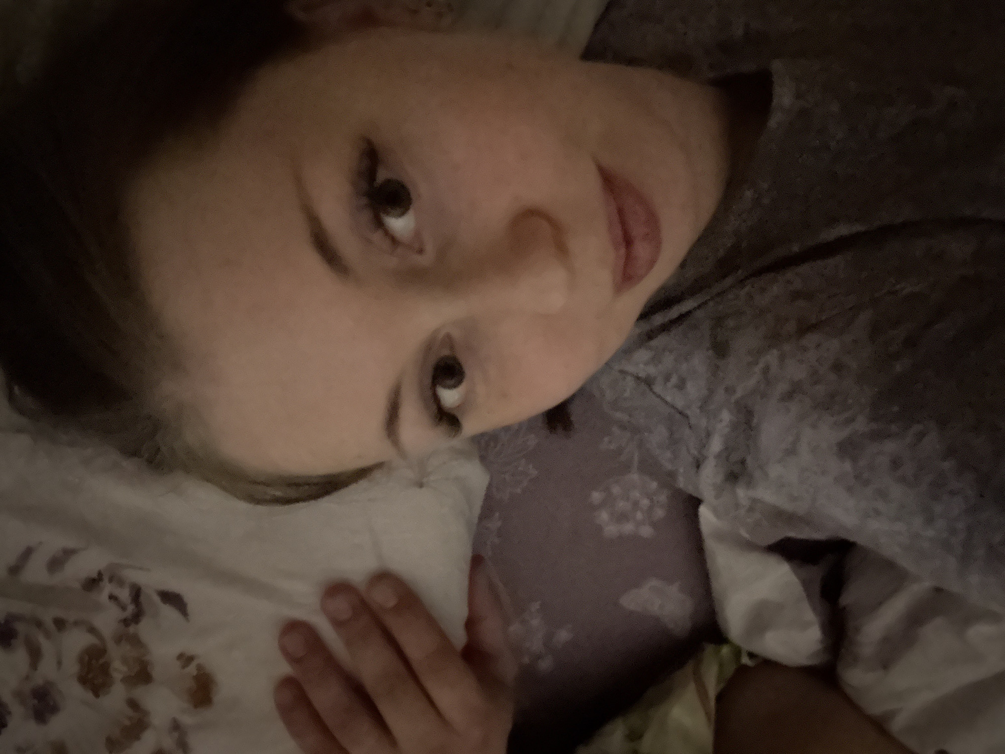 A person with light skin, dark brown hair, and dark eyes is lying down on bedding. The person is wearing a gray shirt. Their right hand is resting on a patterned pillowcase. The background includes pillows and bedding with various floral and abstract patterns. The location is unknown.