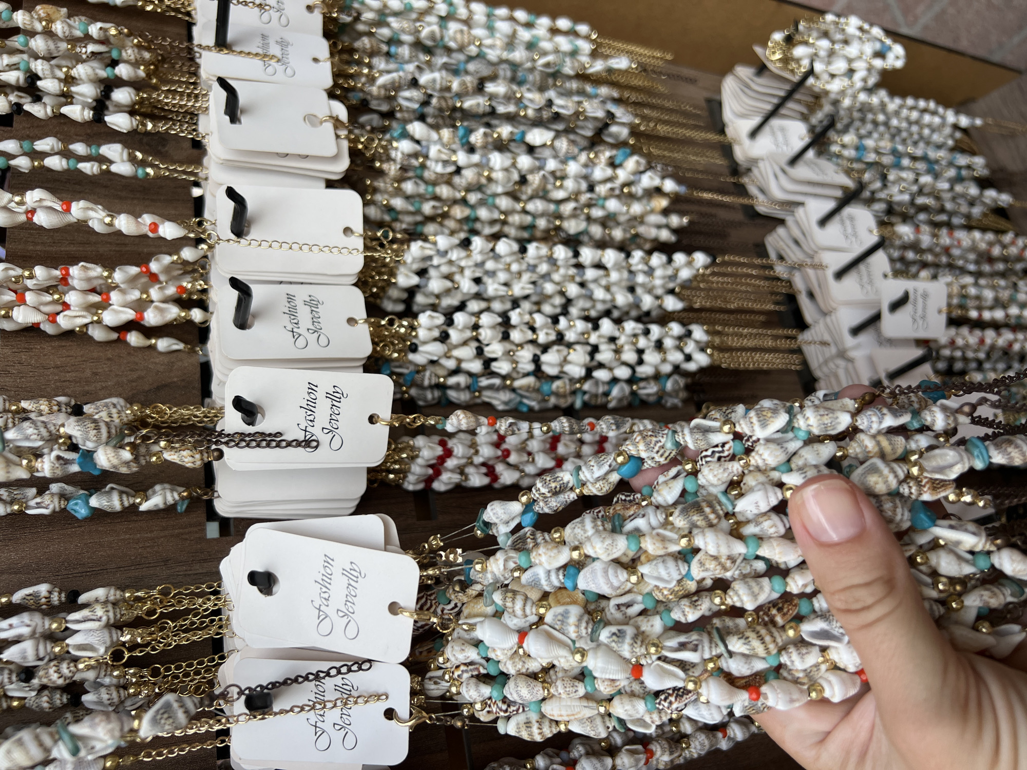 The image shows a close-up view of multiple seashell necklaces and bracelets displayed for sale. The jewelry is made of small, varied seashells, interspersed with gold-colored beads and some turquoise and red accents. Each piece of jewelry is attached to a white tag that reads "Fashion Jeverlly" in a cursive font. The items are hanging from hooks on a wooden display stand. In the foreground, a person's hand is visible, gently touching one of the necklaces. The background is blurred, but appears to be an indoor setting, possibly a market stall or boutique. While the exact location in Alanya, Turkey, cannot be determined from the image alone, the style of the jewelry and display suggests a tourist-oriented shop in a coastal city.
