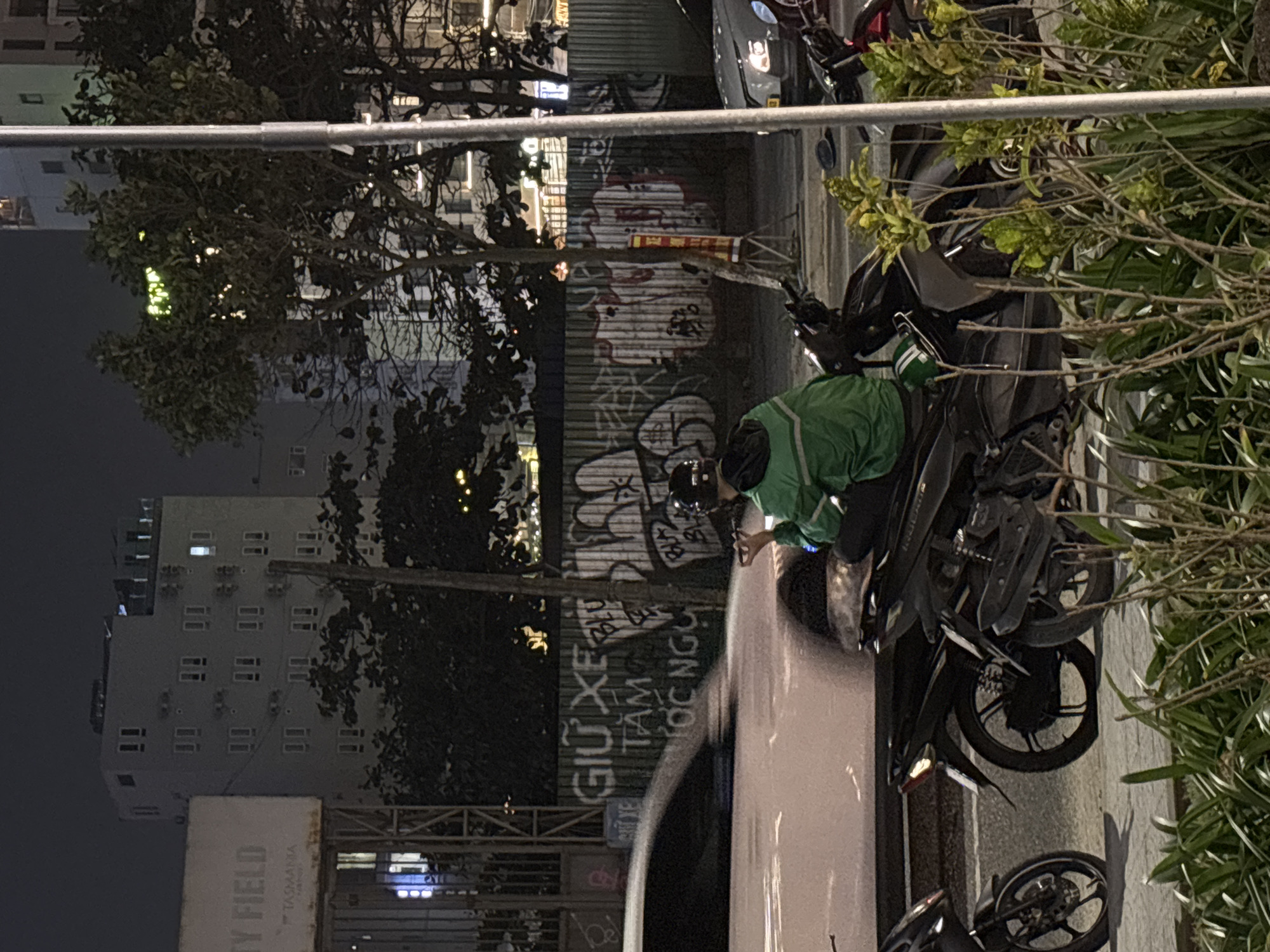 The image depicts a nighttime scene in what appears to be Đà Nẵng, Vietnam. An individual wearing a green jacket and a dark helmet, possibly a motorbike driver, is crouched near a row of parked motorbikes. A wall covered in graffiti serves as a backdrop. The environment includes trees and an urban landscape, with buildings visible in the distance. A sign for "CITY FIELD" is noticeable on one of the buildings. The specific actions are unclear, but the individual seems to be attending to one of the motorbikes.