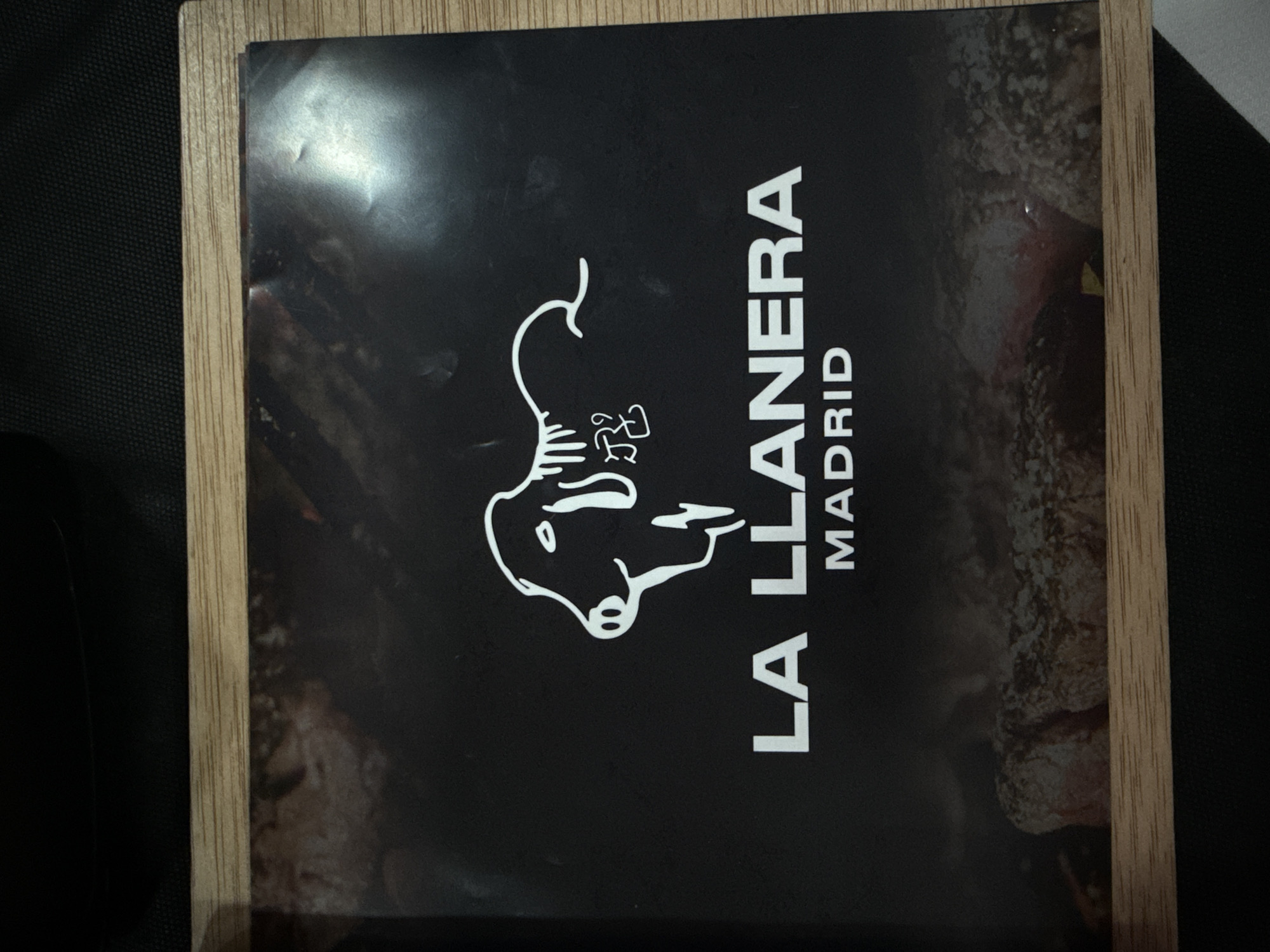 The image shows the cover of a menu for "LA LLANERA MADRID". The cover is black with white text and a white drawing of a bull's head. The bull's head is in the upper center of the cover, and the text "LA LLANERA" is below it, with "MADRID" below that. The background of the cover is a dark, abstract image that appears to be a close-up of meat or a smoky fire. The menu is framed by a light-colored wooden border. There are no people or actions depicted in the image. The location is not visible in the image.