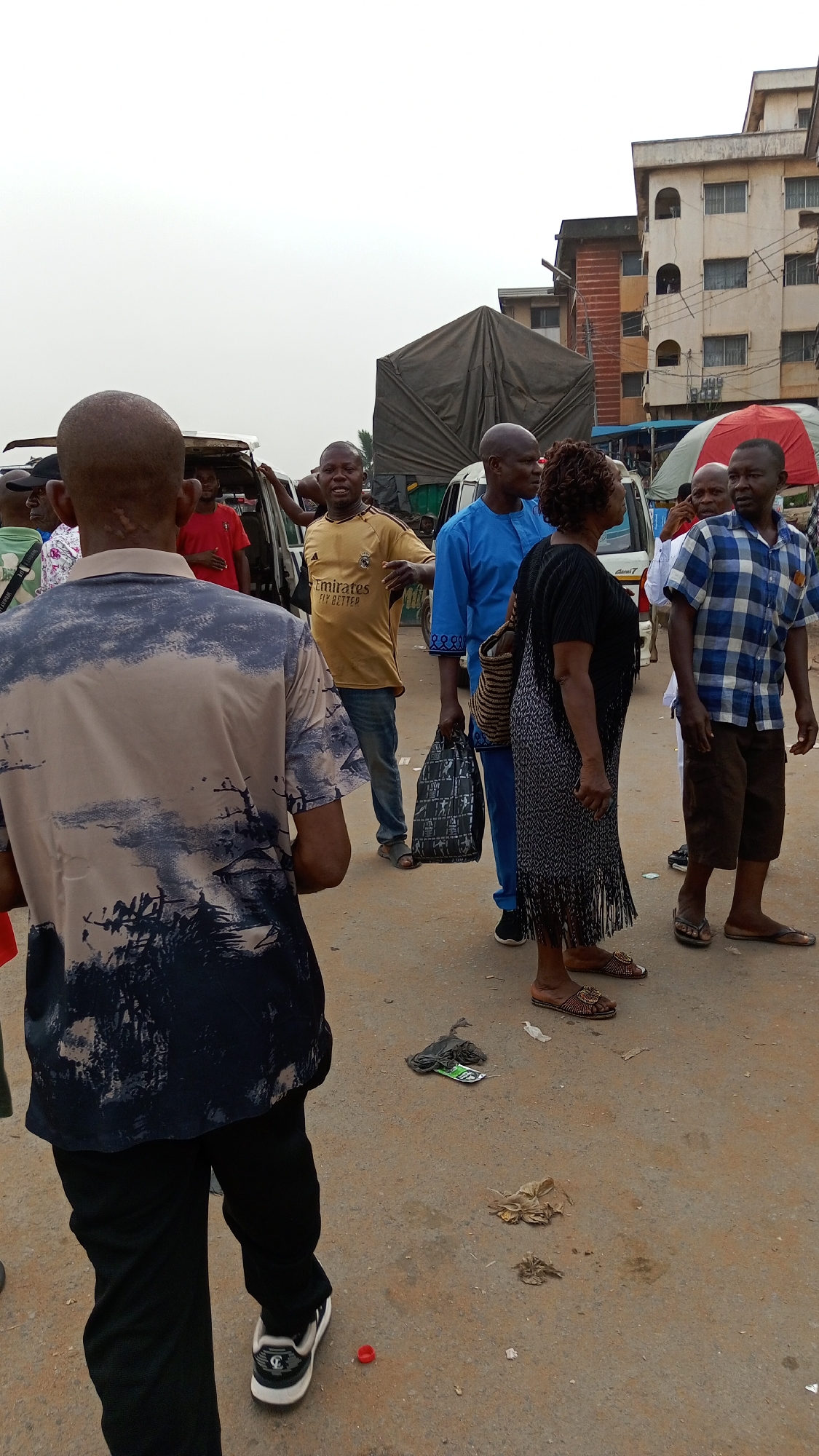 The image captures a street scene in Onitsha, Nigeria. The weather appears overcast. A group of people is gathered, some standing and others near vehicles. The man closest to the foreground has a bald head and wears a patterned shirt. A man in a yellow shirt gestures while another in blue stands beside him. A woman in black is also present. In the background are buildings, parked vehicles, and a covered truck. Litter is scattered on the ground.