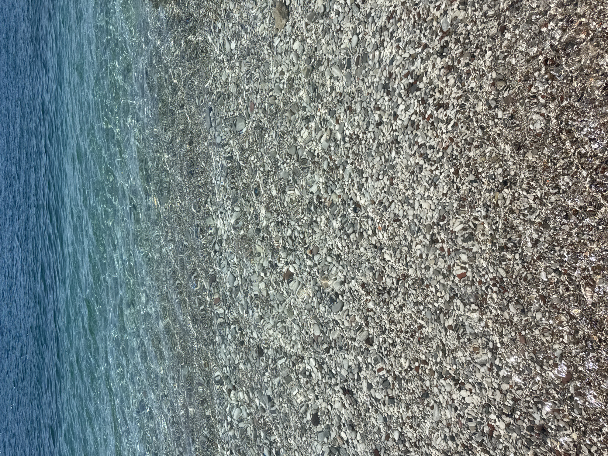 The image depicts a section of the shore in Budva, Montenegro, where the clear, turquoise water meets the rocky beach. The water transitions from a deep blue on the left to a transparent hue closer to the shoreline. The beach itself is composed of small, smooth, gray and white pebbles and stones, visible through the water near the shore. The sunlight reflecting through the water creates patterns and shimmer across the pebbles. No people or animals are visible in the scene.